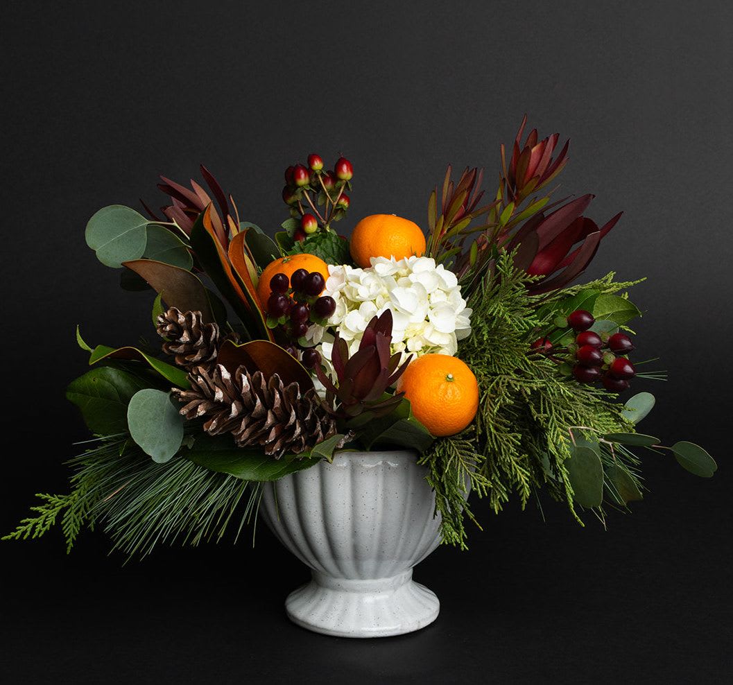 Orangery Collection - Small Compote Arrangement  - An orangery was an elegant conservatory or greenhouse, commonly found on the estates of fashionable northern European residences from the 17th to 19th centuries. These stately structures were designed to shelter orange trees and other delicate fruit plants during the winter months. Oranges held special significance during the holiday season—often given as gifts, tucked into stockings, studded with cloves, or used to infuse festive treats with their bright, citrusy flavor.   This collection blends the freshness of oranges, hydrangea, pine cones and berries with classic holiday greenery—cedar, fir, and magnolia—to evoke the timeless charm and rich tradition of an old-world celebration. ORDER WHILE SUPPLIES LAST! A lovely gift.   Product Details: This arrangement is approximately 10&quot; tall and 10&quot; wide. 