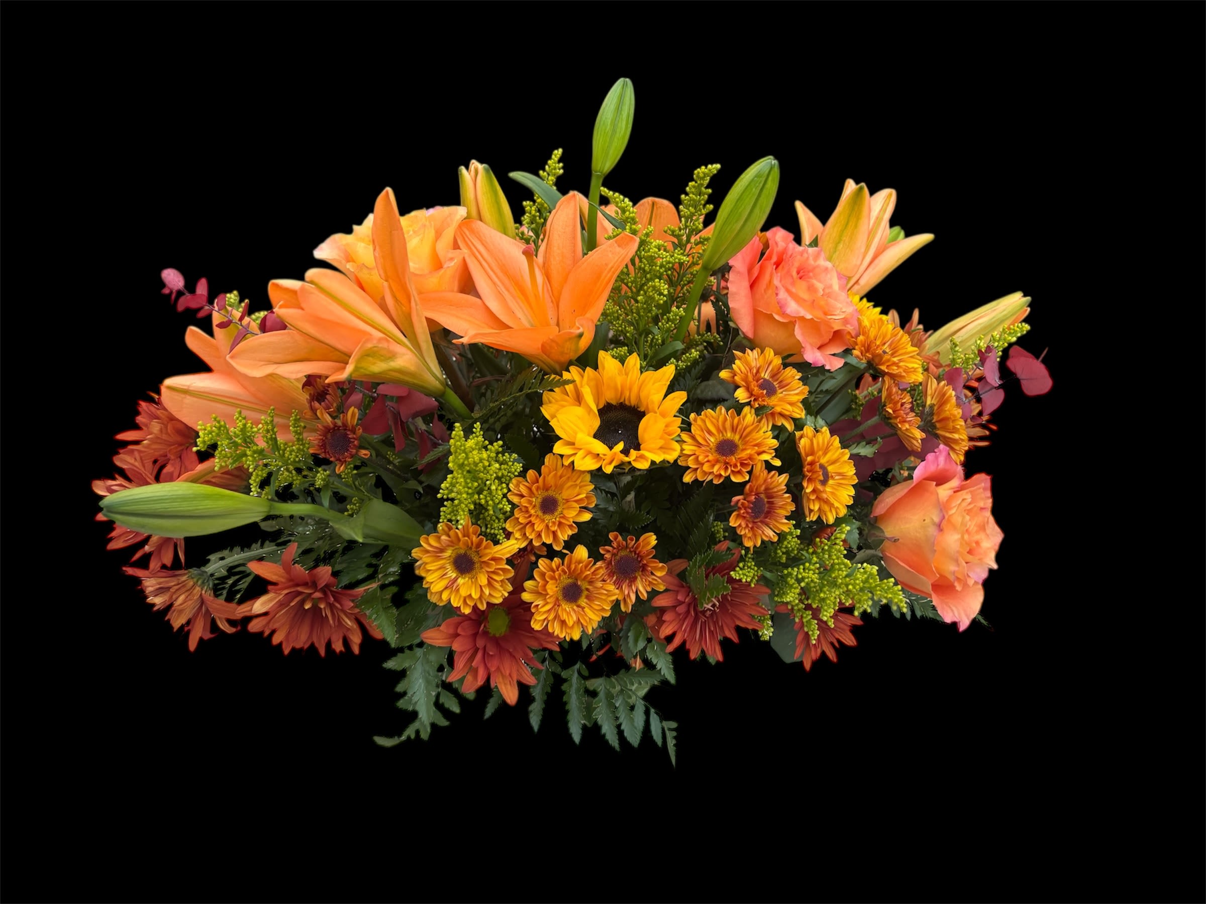 THANKSGIVING CENTERPIECE  - A warm and inviting Thanksgiving centerpiece designed in a low container, featuring vibrant orange lilies, bronze daisies, and airy orange Free Spirit roses. Bold sunflowers add cheerful brightness, while a lush mix of seasonal greenery and golden yellow solidago fills the arrangement with texture and fullness. Rich autumn tones and natural movement make this centerpiece a perfect focal point for any holiday table. 