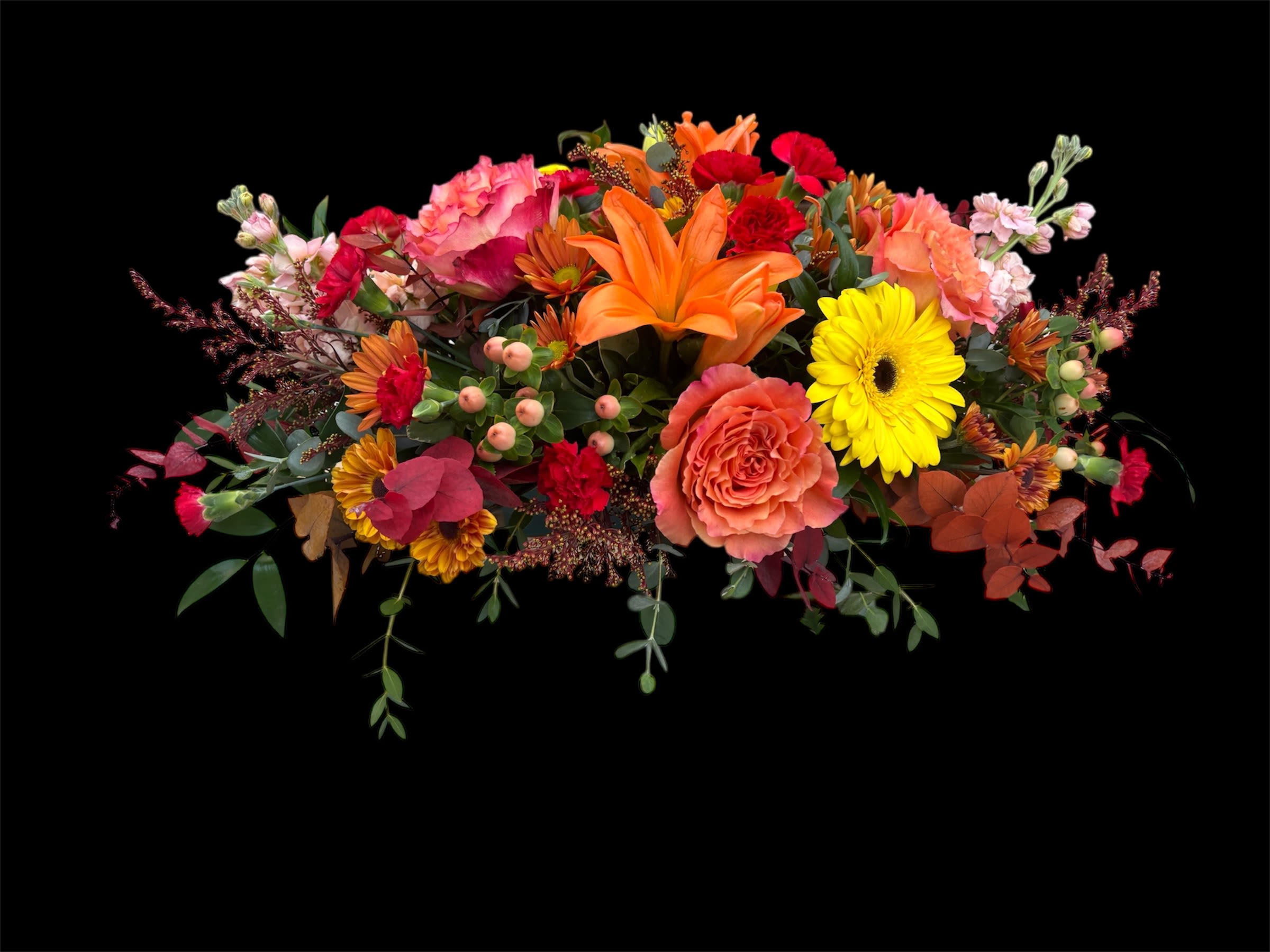 THE AMBER CENTREPIECE  - A warm, autumn-inspired centerpiece featuring vibrant orange lilies and orange Free Spirit roses, accented with cheerful red mini carnations and soft peach hypericum berries. Delicate stock and charming daisies add texture and lightness, while a lush mix of seasonal greeneries brings natural fullness and balance. Rich in color and elegant in style, this arrangement creates a beautiful focal point for any table or celebration. 