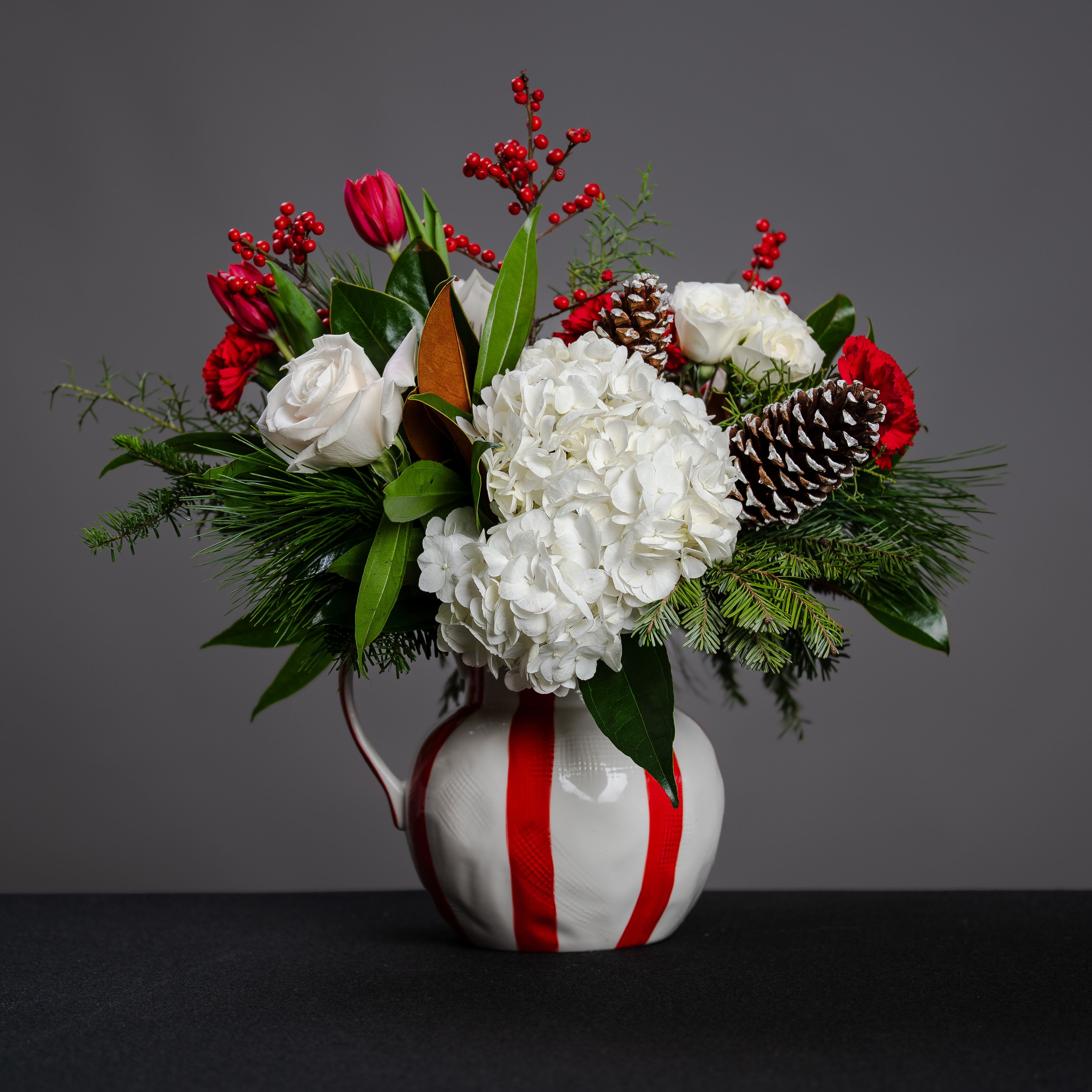 Peppermint Twist - What a gorgeous red and white pitcher!  It's filled with winter greens, hydrangea, roses, spray roses, carnations, and tulips, accented with a snow tipped pine cone. An amazing gift for anyone. The perfect arrangement for the kitchen! Approximate size, 16&quot; tall and 19&quot; wide.