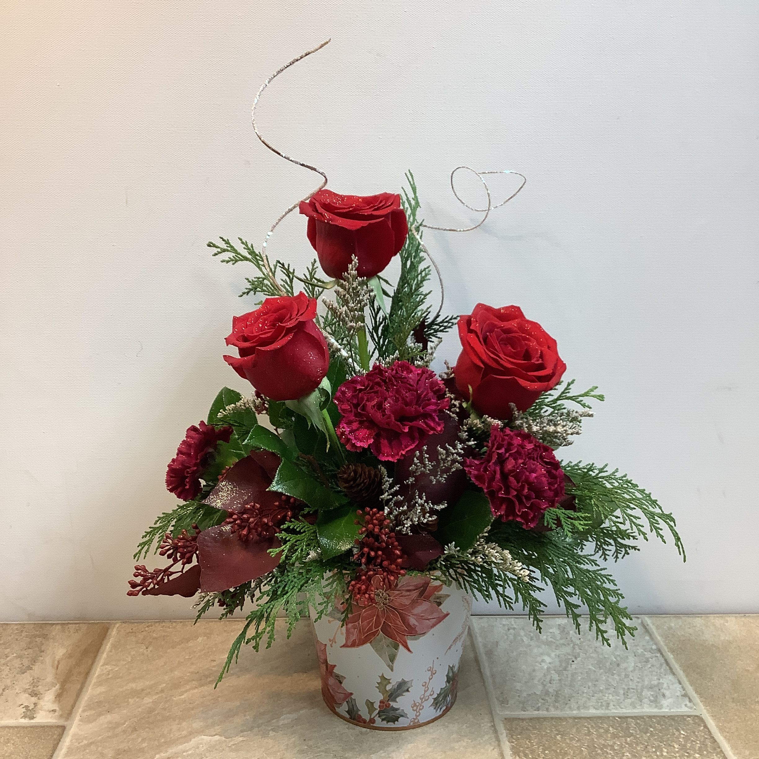 Ruby Delight - This festive bouquet features a rich, traditional holiday palette centered around velvety red roses and deep crimson carnations. The blooms are nestled into a bed of fresh cedar accented with tiny pine cones and clusters of burgundy berries. Delicate frosted accents give the piece a playful sparkling touch. The flowers are arranged in a seasonal metal container decorated with poinsettias and holly, completing the cozy holiday feel.   Primary flowers: rose, carnation