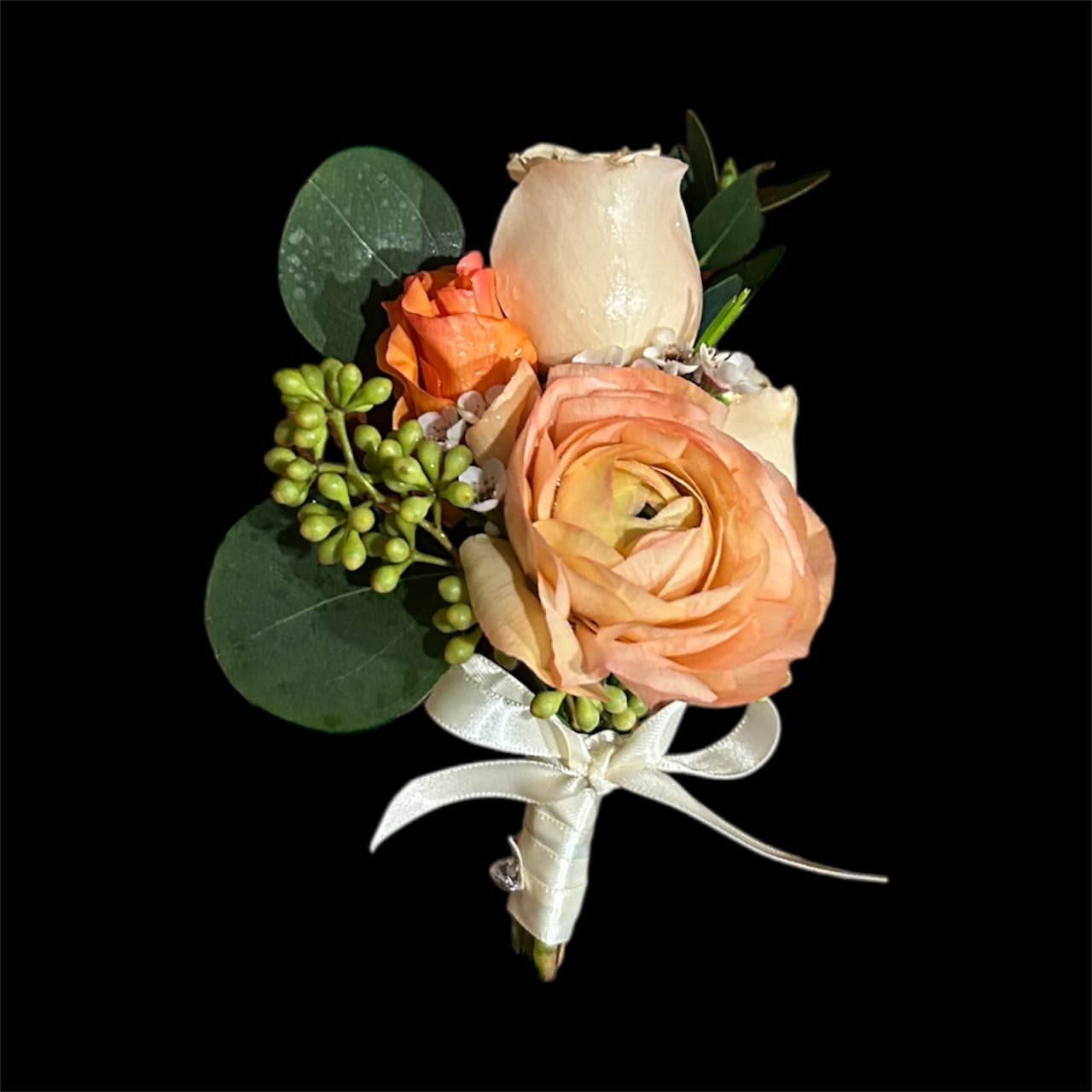 Peach &amp; Coral Charm Boutonniere - A vibrant groom’s boutonniere featuring peach ranunculus, orange star spray rose, and Sahara spray rose, accented with silver dollar eucalyptus, seeded eucalyptus, and parvifolia for a lush, textured look. Finished with a cream-colored ribbon tied in a bow, this design radiates warm, rustic elegance—perfect for romantic or outdoor weddings.