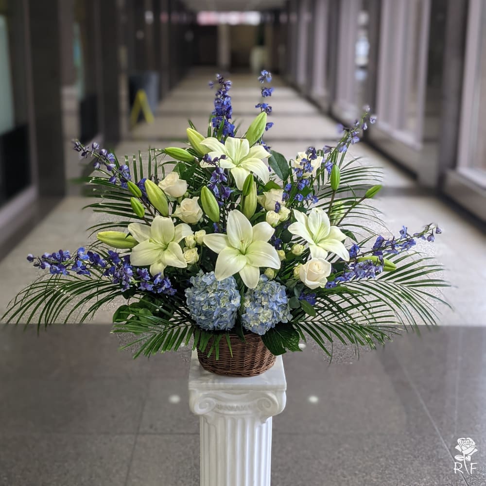 Deepest Condolences  - A blue, white, and green funeral basket for any type of service. Held in a wicker basket, large palms spread out and give a natural spotlight for the feature blooms and colors.
