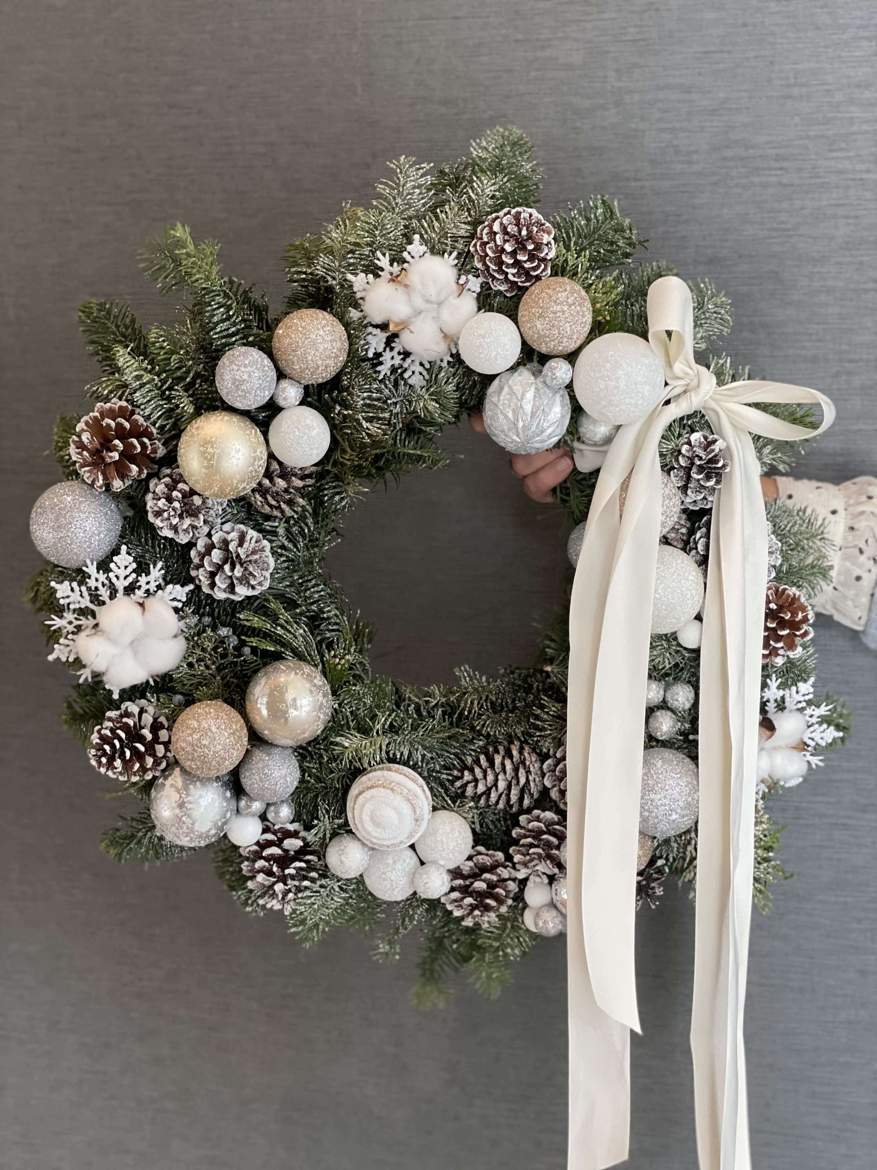 Christmas wreath - This luxurious winter wreath is crafted from fresh evergreens and adorned with a soft palette of snowy white, champagne, and silver ornaments. Frosted pinecones, delicate winter berries, and plush white accents create a magical “winter wonderland” effect. Finished with an elegant long ribbon, this wreath brings a refined, stylish, and festive touch to any space.