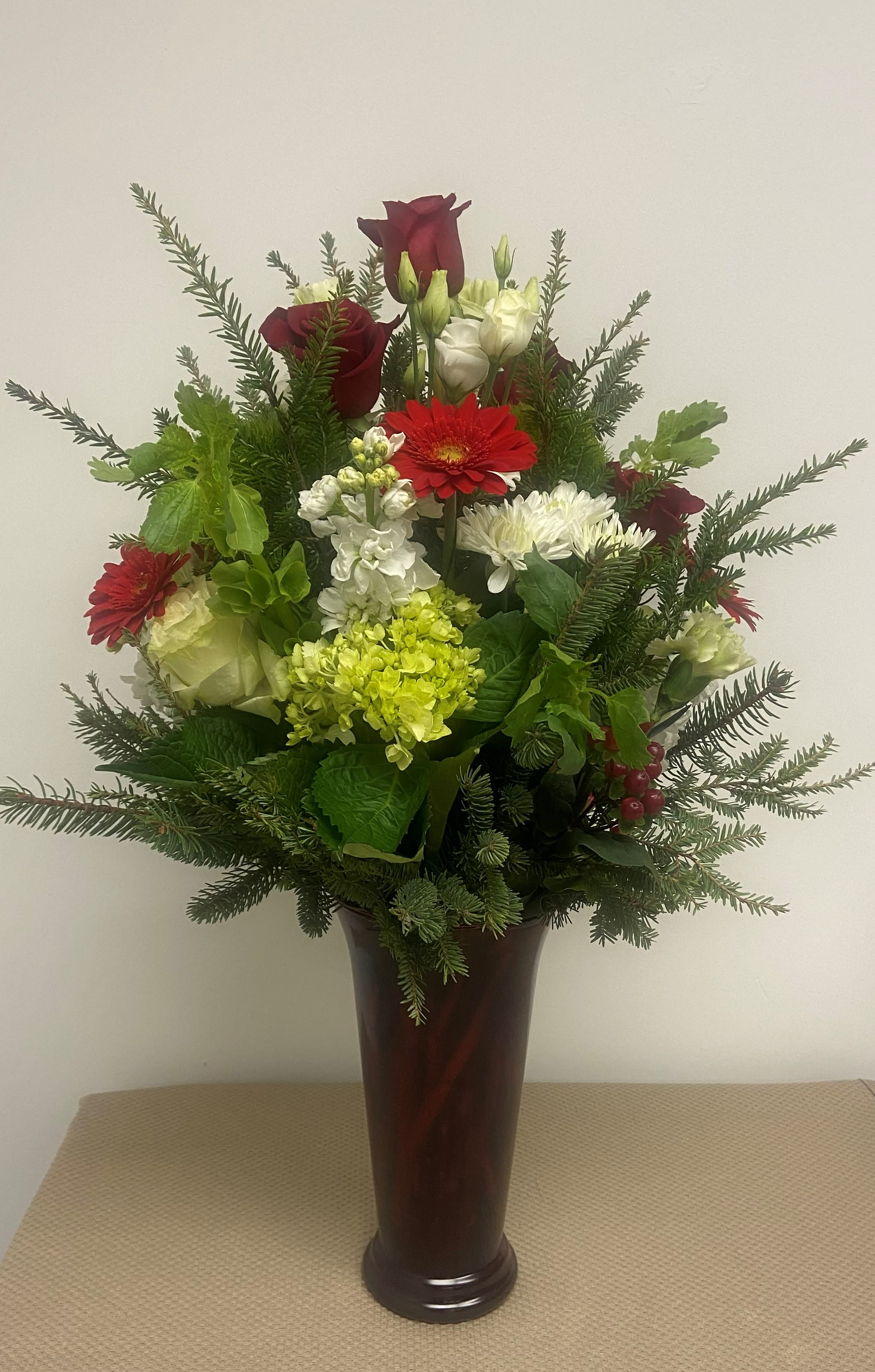 Holiday Beauty - A dramatic collection of designer chosen holiday colored blooms and mixed evergreen foliages, artfully arranged in a tall clear glass vase or pictured vase if available.  A truly colorful expression of holiday bounty and beauty, suitable for all occasions.
