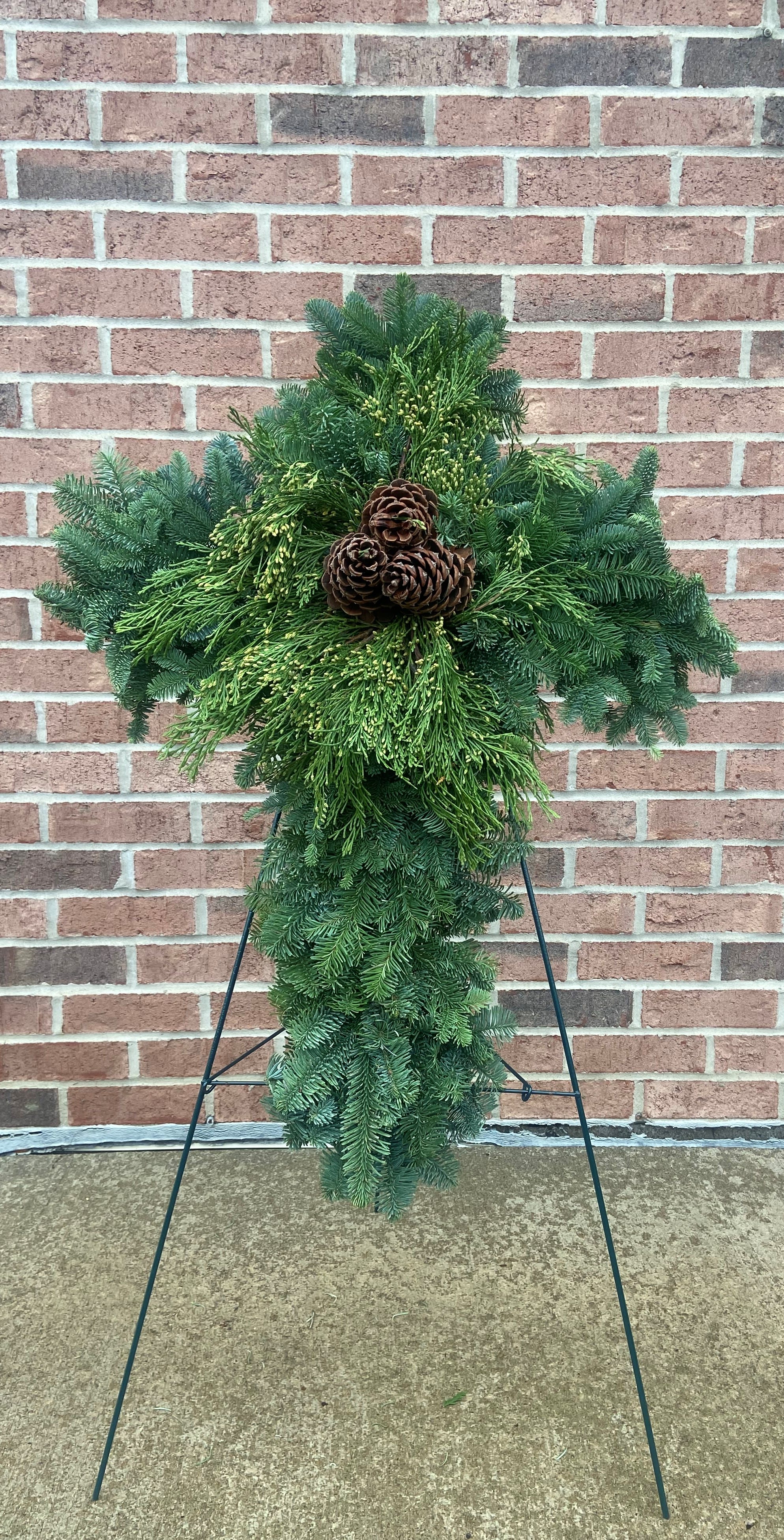 In Remembrance Cross - A lush mixed evergreen cross with a central cluster of pine cones, suitable for sending to the funeral home and then for placement at the cemetery. Cross is approximately 22 inches wide x 32 inches high, tripod is 40 inches high.  Delivery to funeral homes or recipients home only.  Recipient will be responsible for placement at local cemetery.