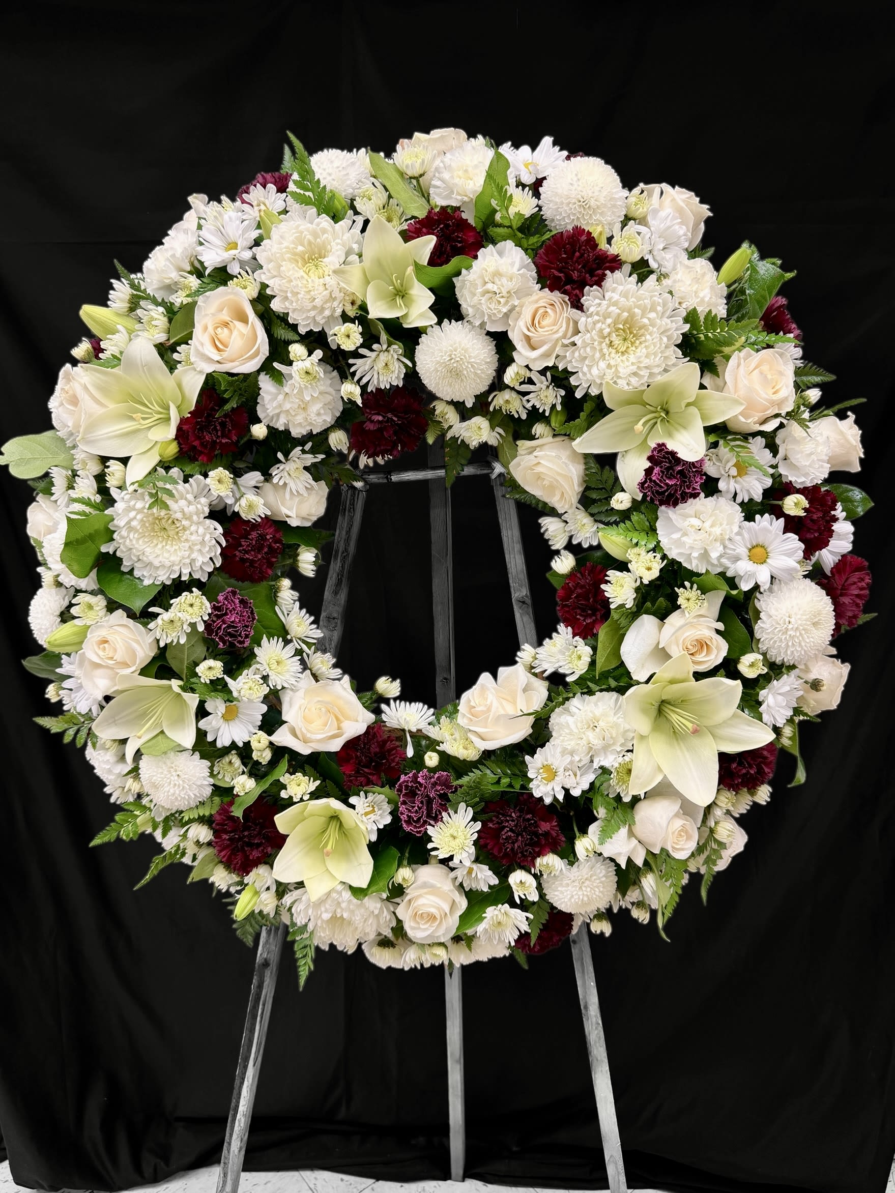 Never - A timeless and graceful tribute in ivory and burgundy. This standing wreath features a delicate blend of white mums, creamy roses, lilies, and rich burgundy carnations—creating a serene and heartfelt expression of love. Designed to honor a cherished life with purity, elegance, and deep respect. 30 Inches