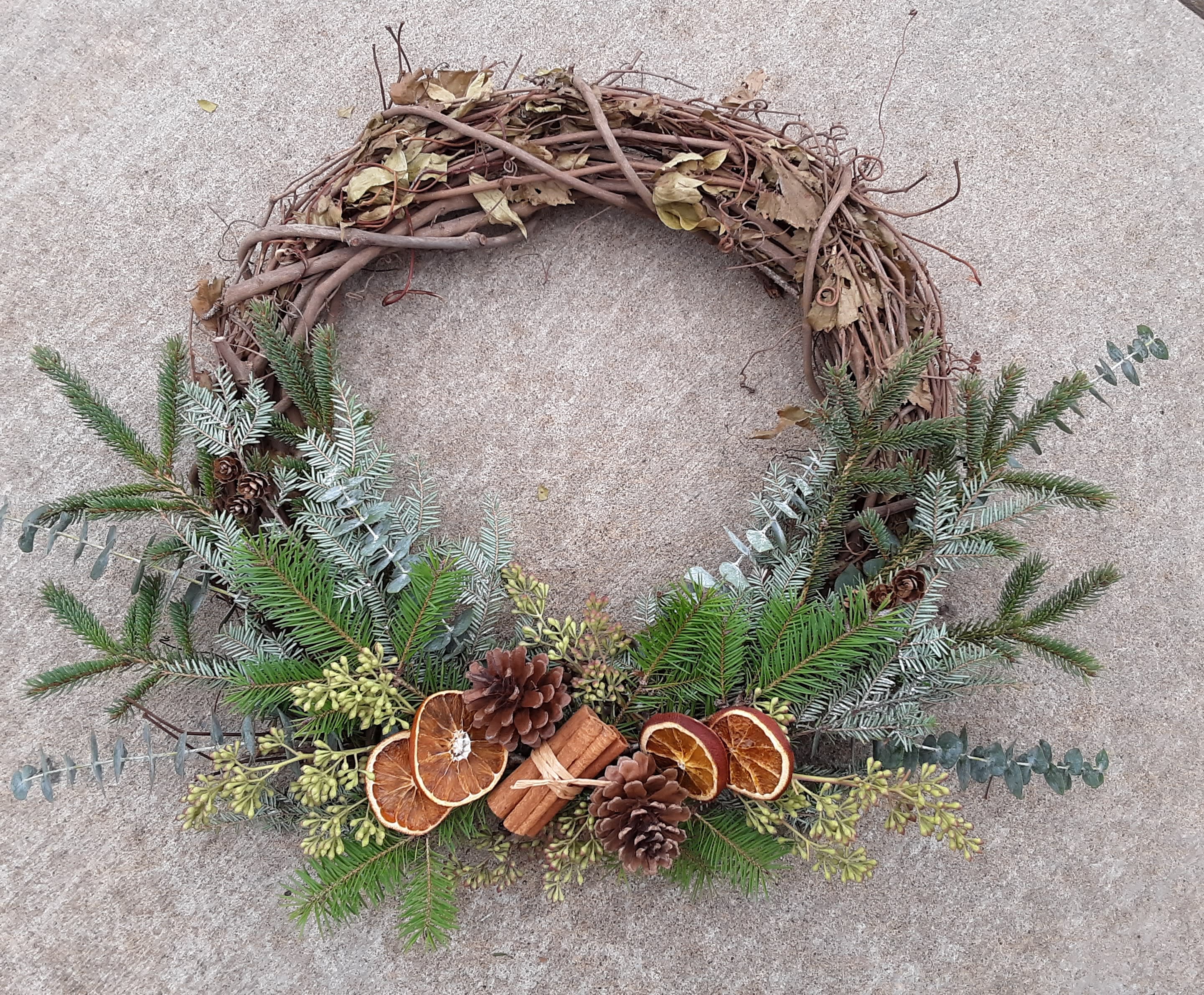 Winter Farmhouse Wreath - This incredible long-lasting rustic grapevine based wreath is handcrafted by our Designer with assorted pines &amp; greens, cinnamon sticks, dried fruits, and other natural elements.  Outside dimensions measure approximately 24" wide and 22 "H on the outside and 12" diameter on the inside.  Perfect for the recipient who loves a natural rustic "farmhouse" look in their decor.   Suitable for indoor or outdoor presentation.  Upgrade includes a tripod for a free standing presentation.