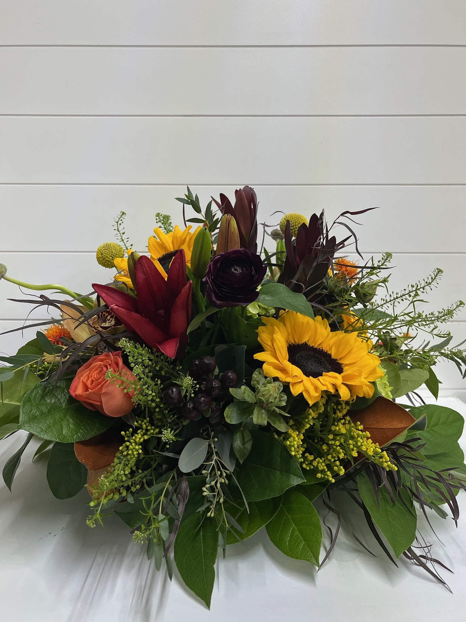 Golden Garnet - Golden Garnet – Thanksgiving &amp; Autumn Centerpiece  Celebrate the warmth of the season with Golden Garnet, a rich and radiant autumn centerpiece designed to elevate your Thanksgiving table. This stunning arrangement features sunflowers, roses, lilies, billy balls, and other seasonal blooms, artfully arranged to capture the heart of fall.  A luxurious palette of burgundy, orange, and golden yellow brings both depth and glow—perfect for creating a welcoming atmosphere for family gatherings and holiday meals. Whether placed at the center of your feast or given as a festive gift, Golden Garnet brings the essence of autumn’s beauty to every moment.  Flowers and colors may vary depending on availability.