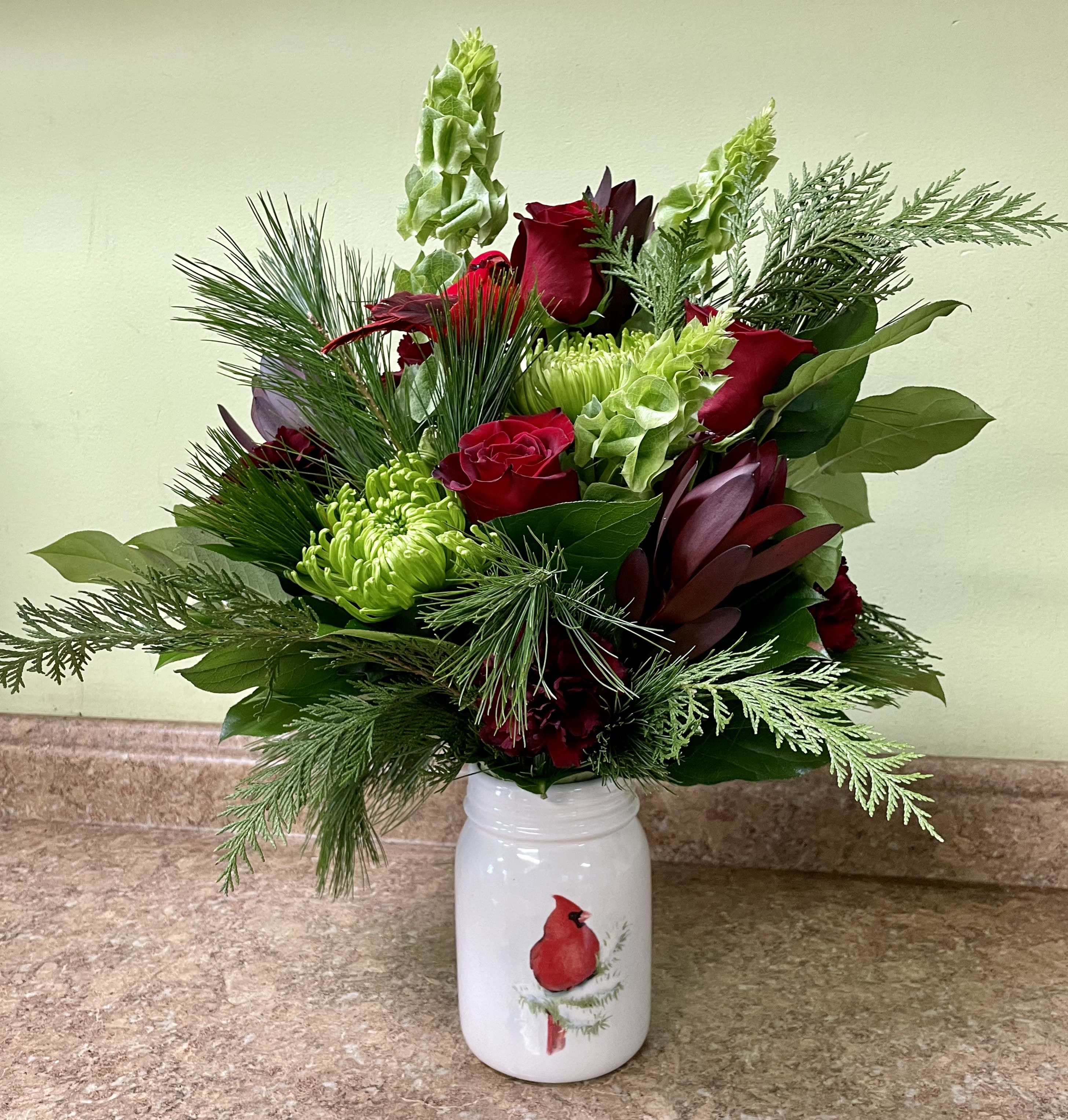 Winter Cardinal - A winter classic....beautiful, fragrant greenery and seasonal fresh flowers combine with a cardinal to fill this keepsake ceramic mason jar. A perfect, thoughtful gift for the holidays. Approximately 20&quot;H X 12&quot;W. Deluxe and premium are made a little fuller with greenery and have more roses added.