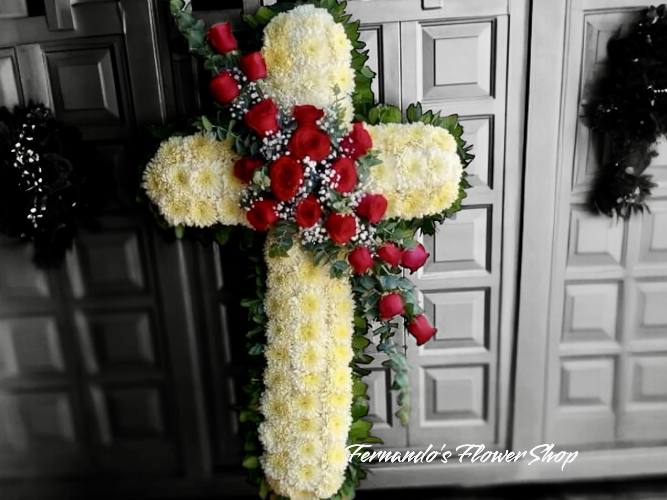 Angel’s Embrance  - This is a beautiful floral cross arrangement made with densely packed white chrysanthemums, symbolizing peace and eternal rest. The cross is accented with a striking cluster of vibrant red roses at the center, representing love and remembrance. Lush green foliage outlines the cross, adding contrast and depth. This elegant design serves as a heartfelt tribute for a memorial or funeral service. The bottom of the image shows the text “Fernando’s Flower Shop,” indicating the florist.