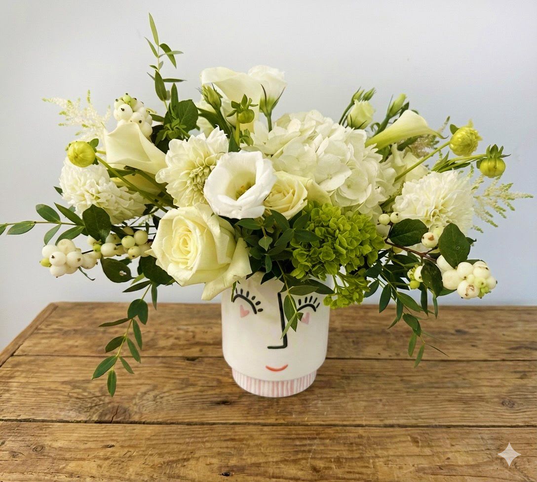 Smiling White - Bright, whimsical, and full of charm — Smiling White features a luxurious mix of creamy roses, white hydrangeas, calla lilies, dahlias, snowberries, and fresh greens. Arranged in a cheerful face vase that beams personality, this design is perfect for sending joy, gratitude, or simply brightening your space with a soft, modern twist.  Details: 	•	Premium blooms: white roses, hydrangeas, calla lilies, dahlias, snowberries 	•	Presented in a playful ceramic smiley face vase 	•	Approx. size: 10-11” W x 10” H 	•	Complimentary gift message card included