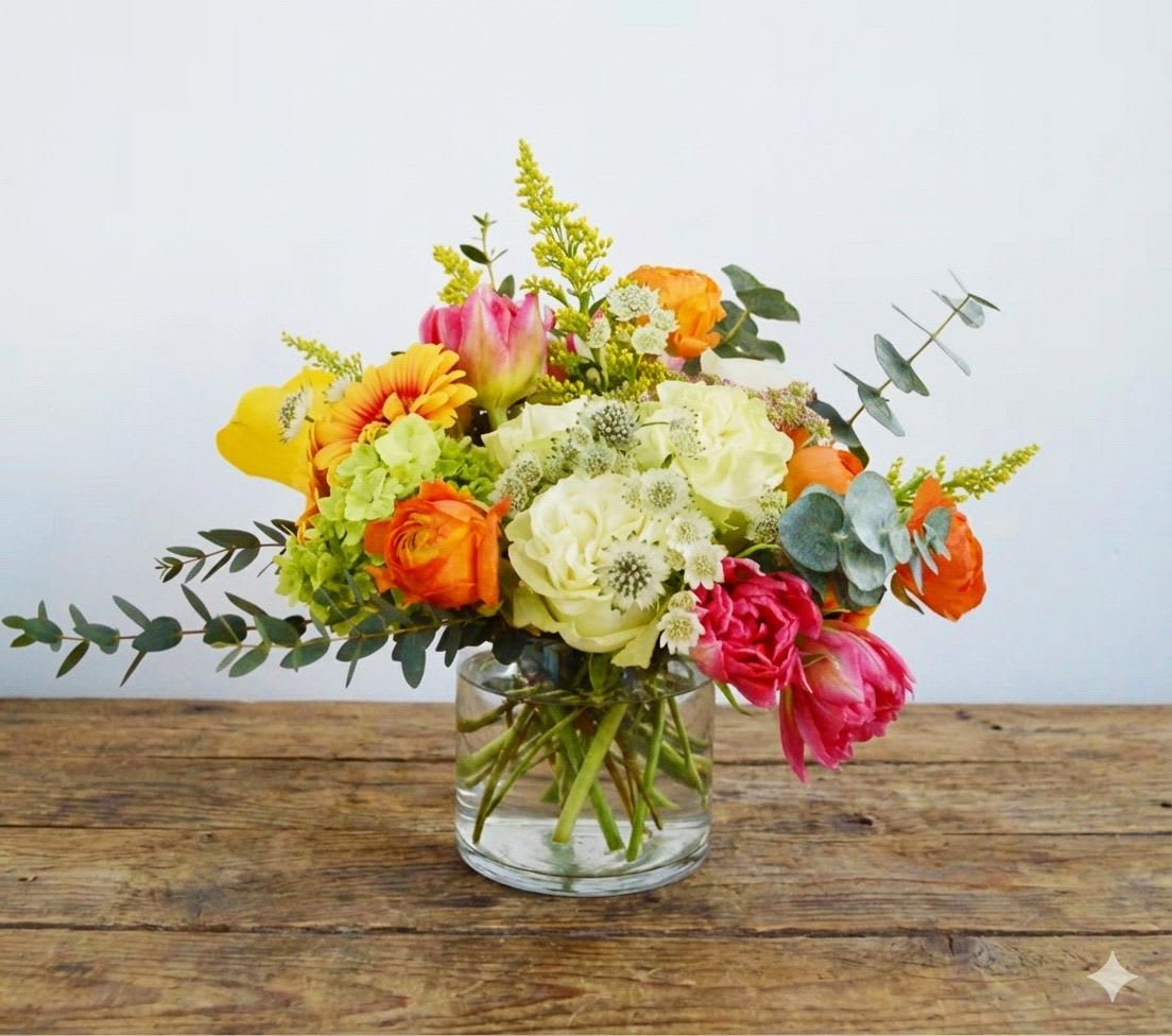 Playground  - This arrangement is presented in a pinched glass vase and accented by bright and cheerful color tones. Flowers include white roses, white calla lilies, orange free spirit roses, green hydrangea, tulip and assorted fillers and greens. Standard size pictured. Larger sizes available.