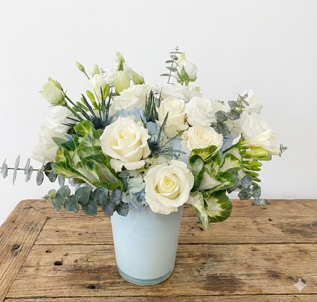 Ocean Breeze - Drift into calm with Ocean Breeze, a serene floral arrangement inspired by coastal air and open skies. Featuring creamy white roses, soft blue hydrangeas, lisianthus, and delicate thistle accents, this bouquet evokes the gentle rhythm of waves and salt-kissed air. Lush and variegated greens add natural texture, all arranged in a sleek white or clear vase for a fresh, contemporary finish.  Perfect for sympathy, thank-you gifts, housewarmings, or any moment that calls for peaceful beauty.  Details: 	•	Premium blooms: white roses, lisianthus, blue hydrangea, eryngium (thistle), eucalyptus 	•	Arranged in a modern white ceramic vase 	•	Approximate size: 10–11” wide 	•	Complimentary message card included  Care Tips: Keep in a cool location, away from direct sunlight. Refresh water daily and trim stems every few days for long-lasting freshness.