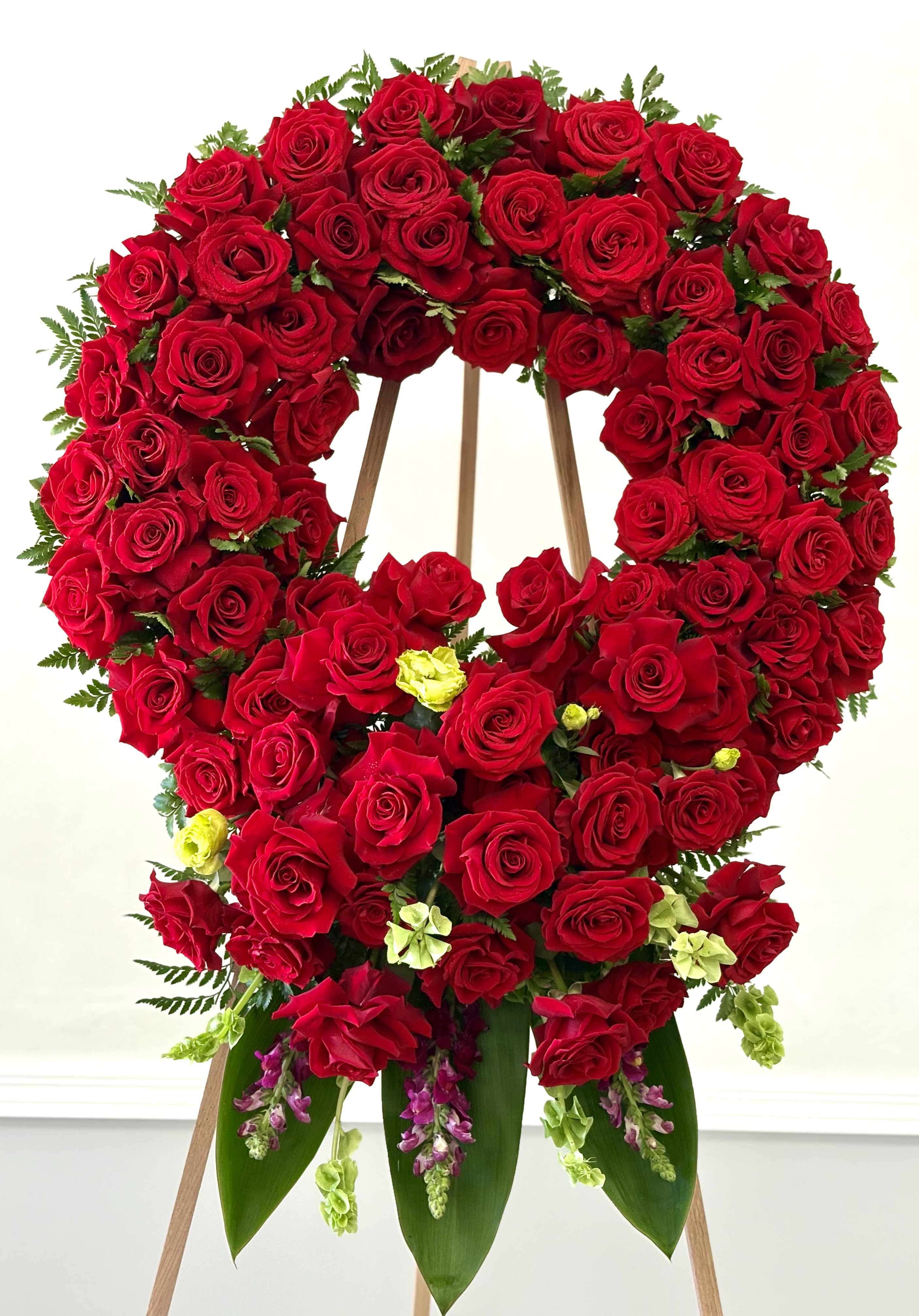 Eternal Red Tribute By VLM Flowers - A stunning wreath overflowing with premium red roses, artfully accented with touches of soft green lisianthus and cascading snapdragons. Lush greenery frames the vibrant blooms, creating a bold and elegant expression of love, respect, and remembrance. This timeless design offers a heartfelt tribute that speaks with beauty when words fall short.