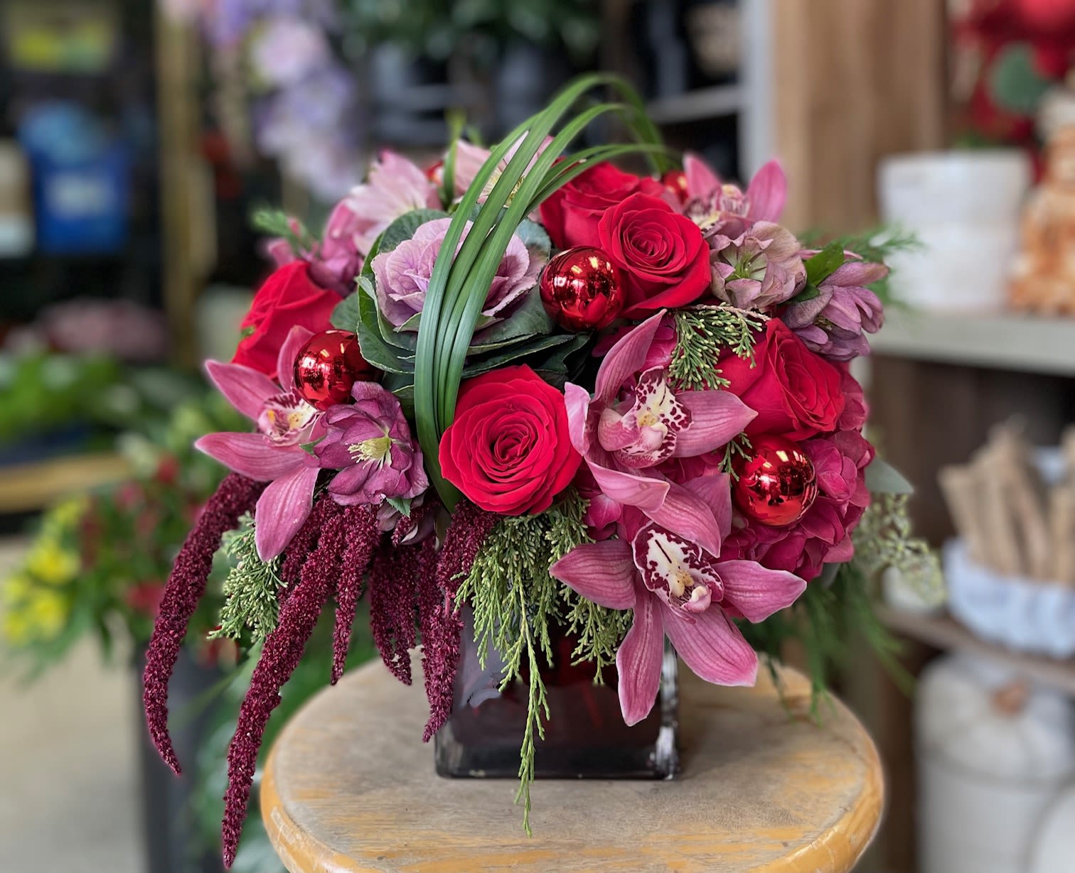 The Velvet Noel - Bold and sophisticated, Crimson Luxe captures the magic of the season with velvety red roses, lush red hydrangeas, exotic cymbidium orchids, and rich purple kale. Cascading red amaranthus and festive ornament accents add drama, while helleborus and fresh evergreens bring seasonal charm. Arranged in a modern red cube, this showstopping piece is perfect for an elegant holiday centerpiece or an unforgettable gift.