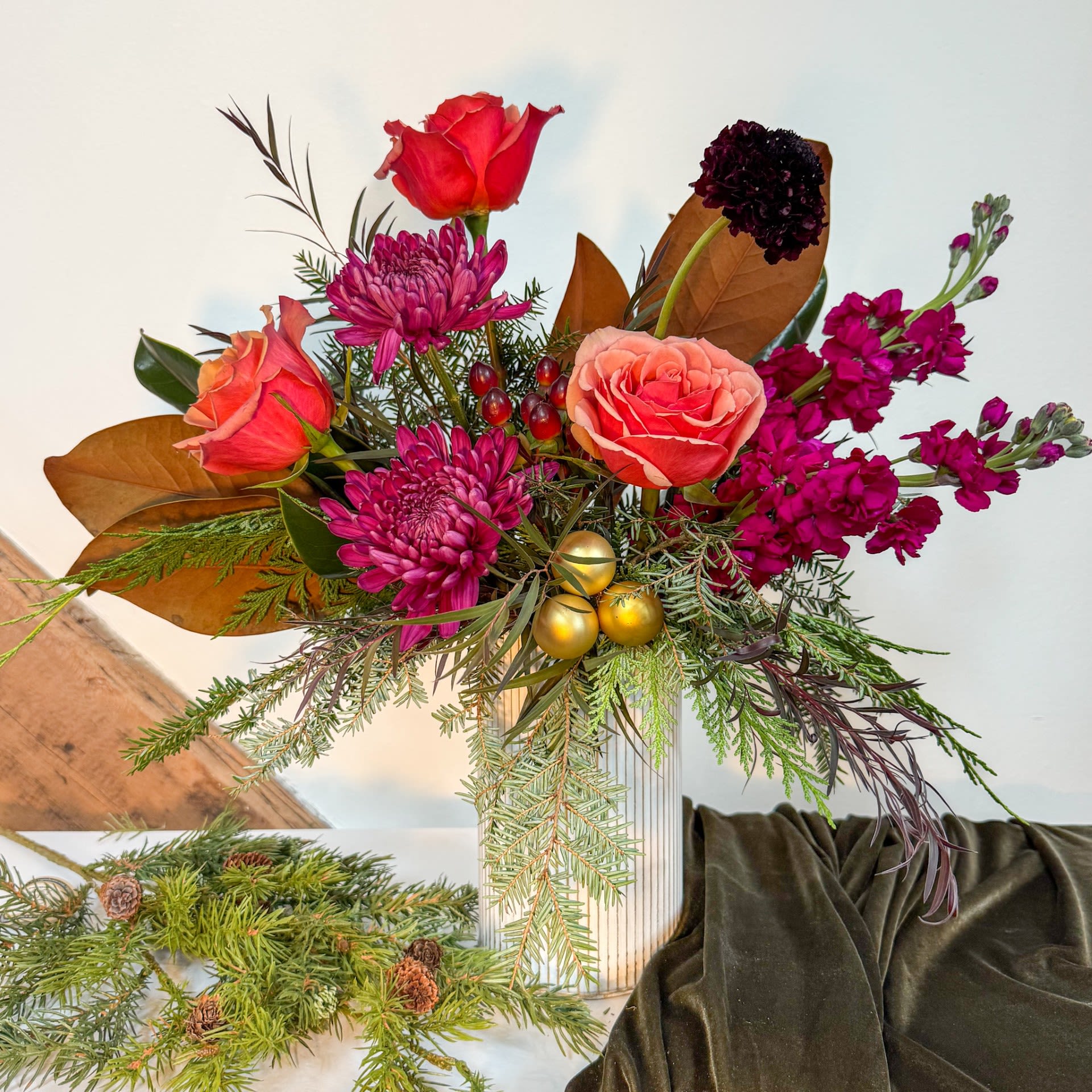 Magnolia Glow - Celebrate the season with Magnolia Glow. A luxurious blend of magnolia leaves, roses, rich purple mums, and dark scabiosa. Golden holiday ornaments add a festive touch. Measure approx 10&quot; x 14&quot;