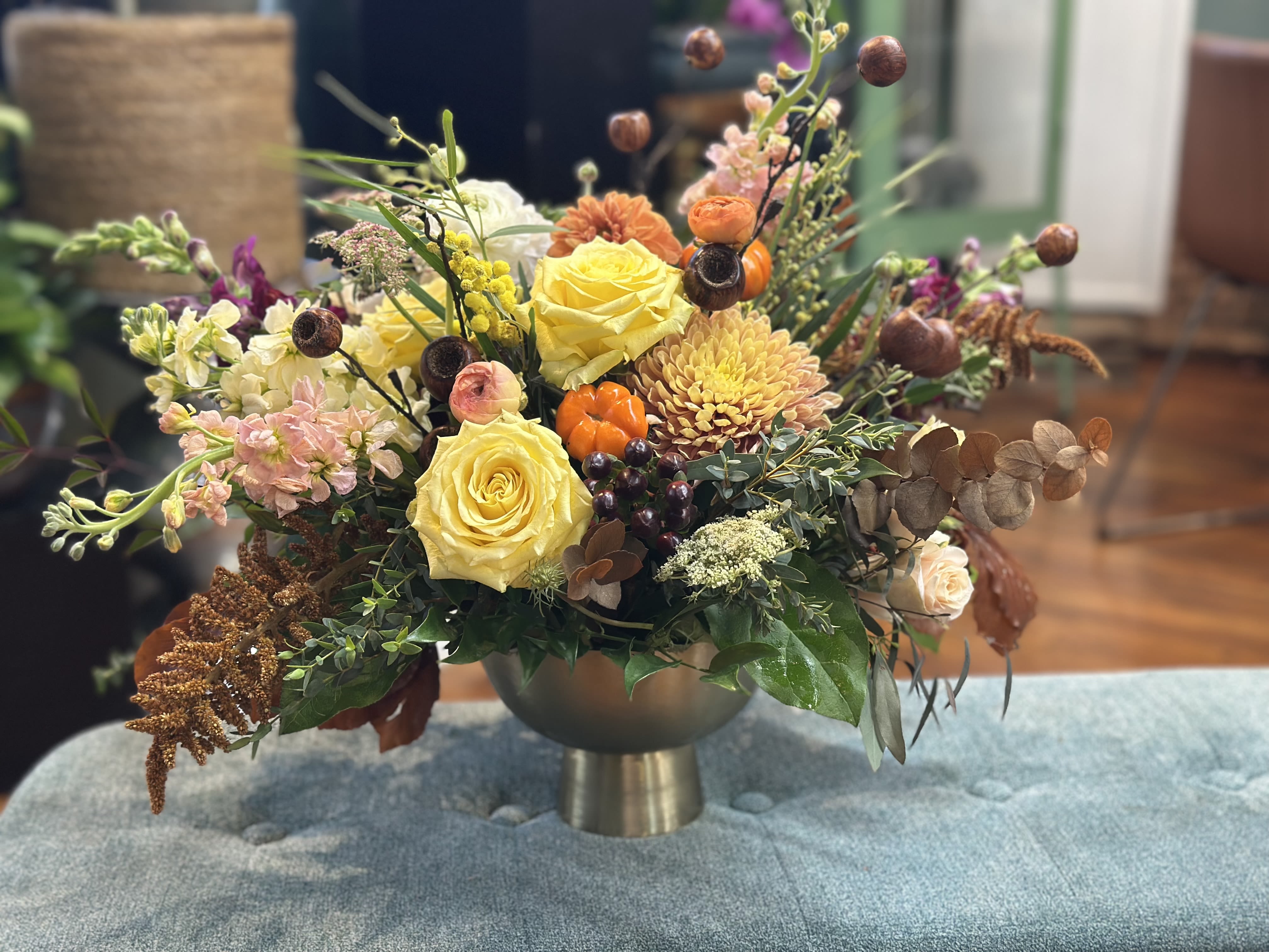 Autumn Reverie - Bring the warmth of the harvest to your table with Autumn Reverie. Roses, dahlias, and chrysanthemums in golden yellow, soft cream, blush pink, and warm orange spill playfully from a gold pedestal vase, layered with seasonal textures and visual intrigue. This centerpiece glows with the spirit of autumn — perfect for Thanksgiving tables, family gatherings, or celebrating the season’s bountiful beauty.