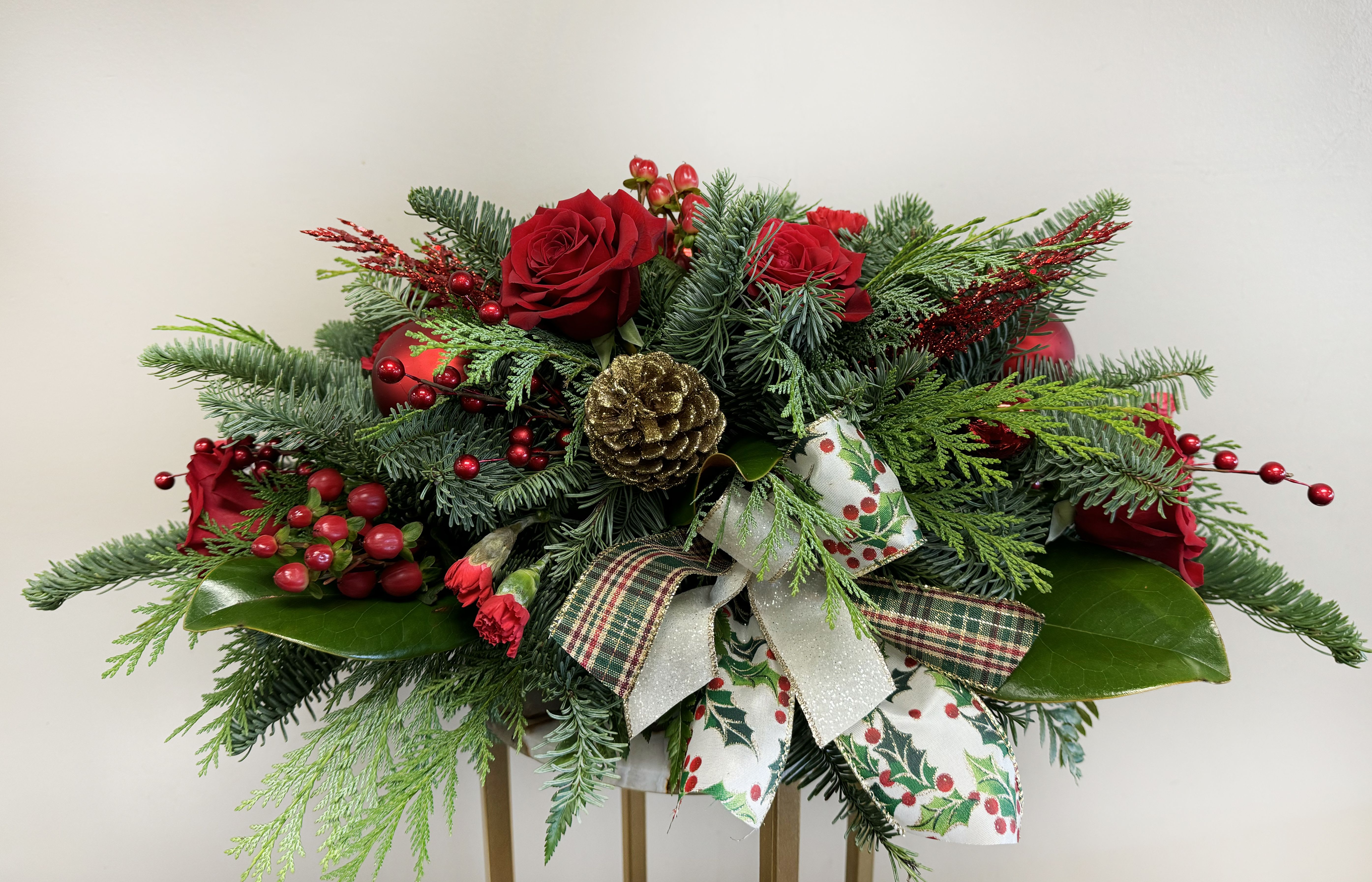 Classic Christmas Centerpiece - Add timeless elegance to your holiday celebrations with our Classic Christmas Centerpiece. Featuring lush noble fir boughs, cedar, magnolia leaves, six vibrant red roses, red mini carnations, red hypericum berries and festive accents like berries, gold pinecones, and decorative ribbons, this arrangement is perfect for your dining table or mantel. Its rich colors and holiday charm make it an ideal choice for spreading seasonal cheer or gifting to loved ones. Approximately 6&quot; wide by 12&quot; long. 