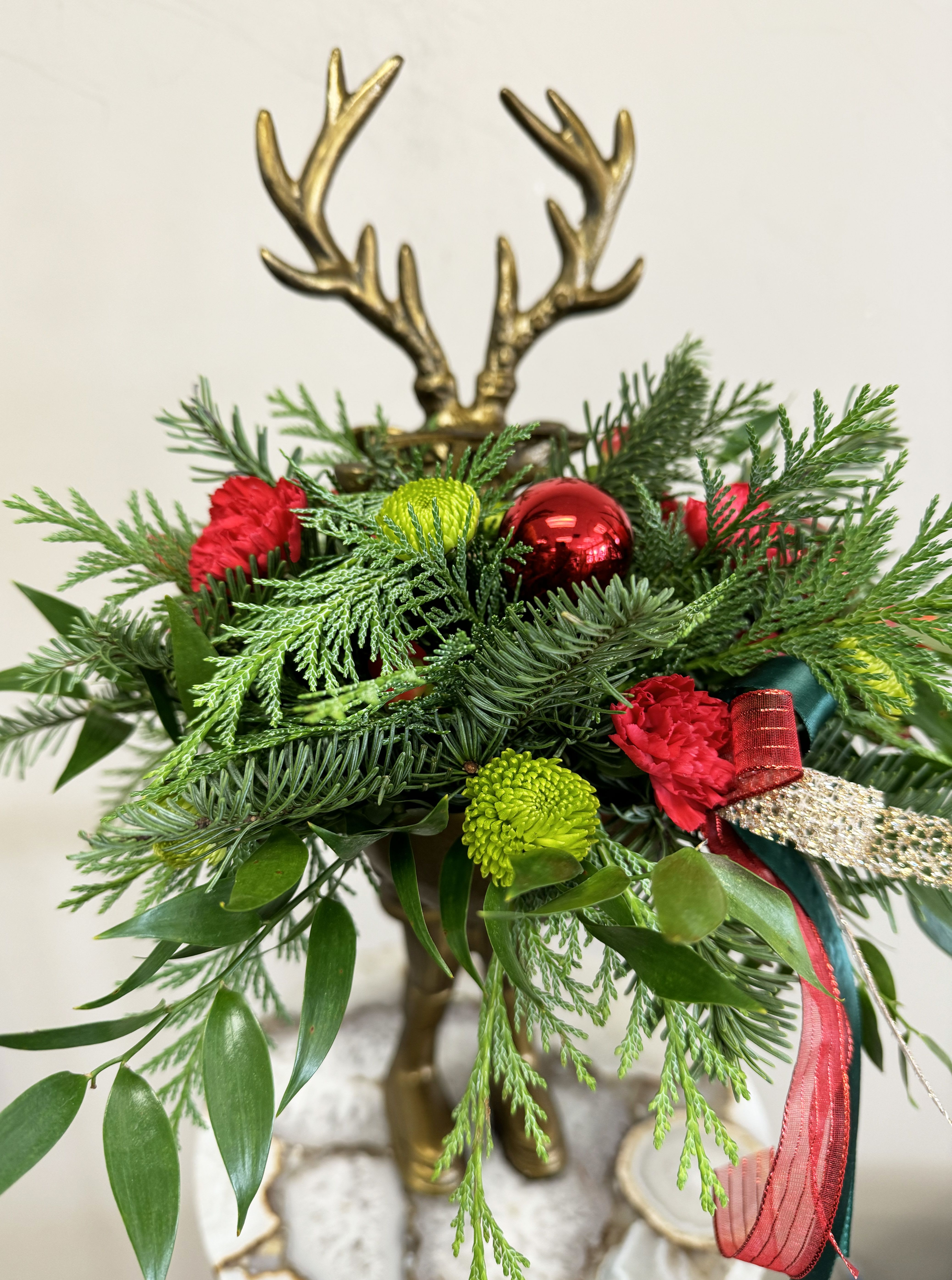 Whimsical Woodland Reindeer  - Showcased in a charming keepsake brass reindeer vase, this festive display features lush seasonal greenery, vibrant mini red carnations, and cheerful green button mums, accented with sparkling ornaments and elegant ribbons. Perfect as a holiday centerpiece, or unique gift, this arrangement adds a touch of rustic charm and holiday cheer to any space.
