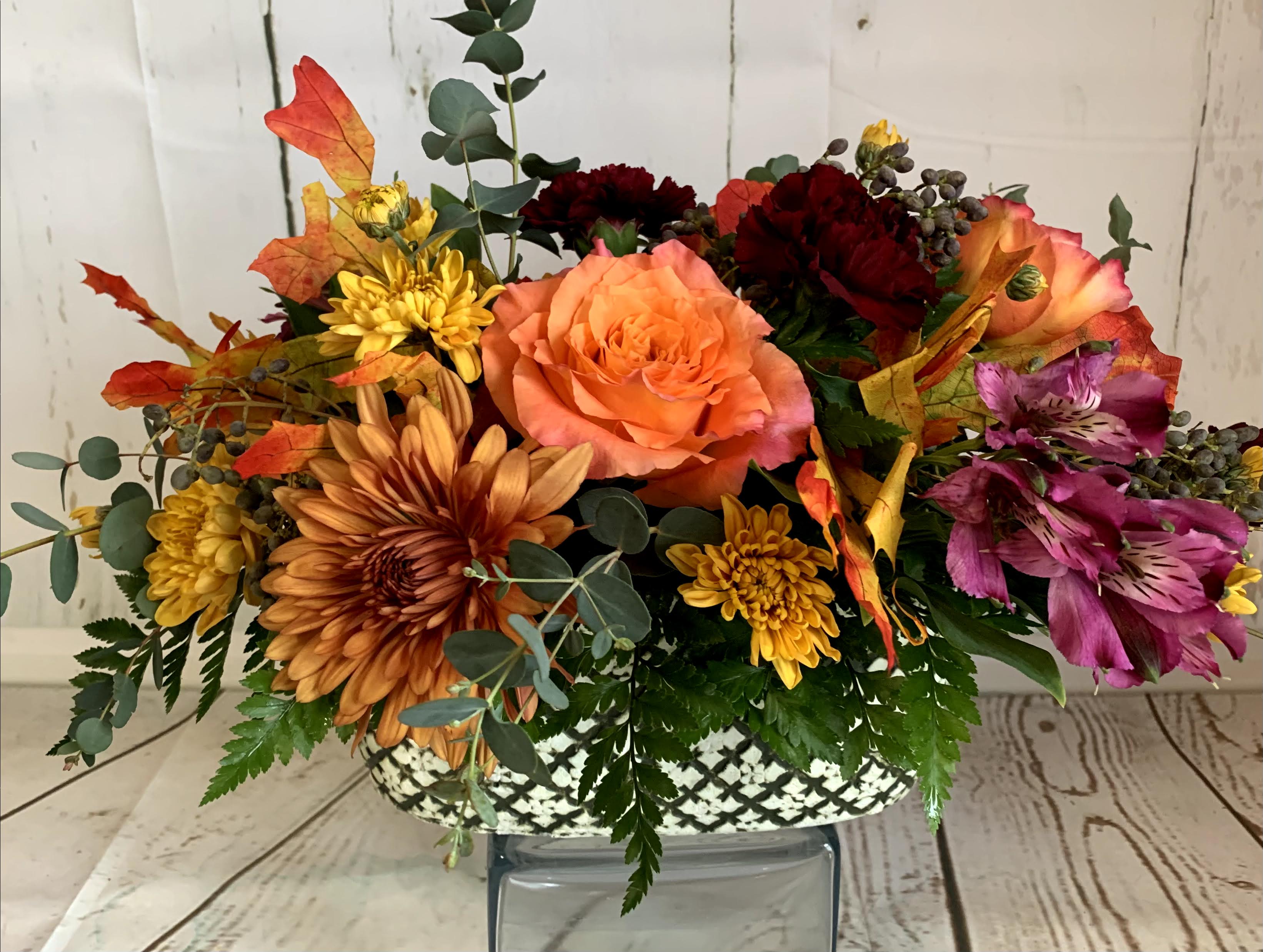 Thanksgiving Centrepiece - Our Thanksgiving Centrepiece Arrangement is a vibrant celebration of the season's rich colors and natural beauty. Featuring an abundant mix of autumnal blooms with shades of gold, deep burgundy, and warm oranges, this arrangement is accented with lush greenery, fall leaves, and seasonal elements. Set in an elegant, low-profile vase or rustic wooden container, the centrepiece is designed to complement your Thanksgiving table without obstructing conversation. The warm hues and natural textures create a cozy, welcoming atmosphere, making it the perfect finishing touch for your holiday gathering.