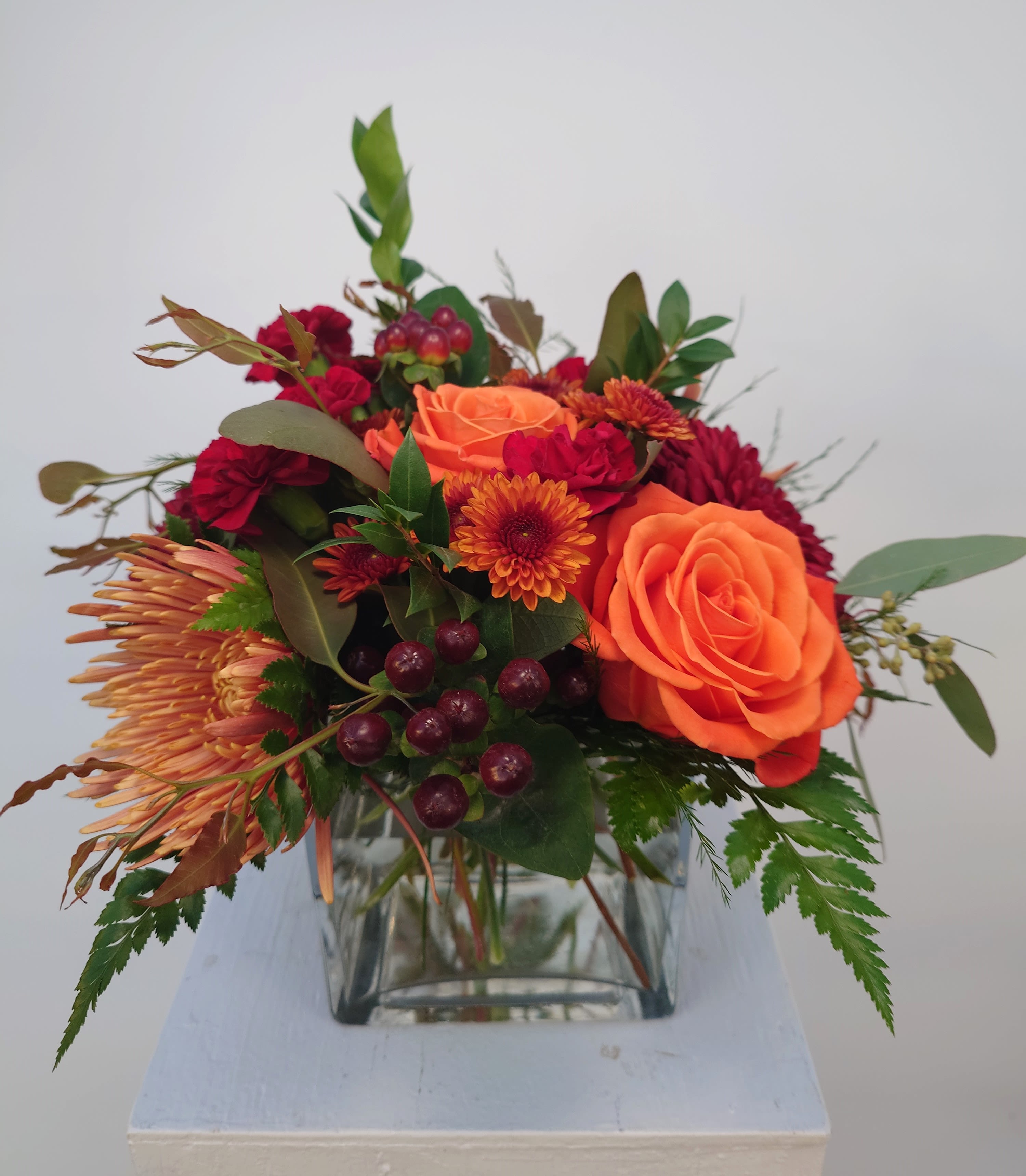 Seneca Sunset Centrepiece - Inspired by the breathtaking colors of a Finger Lakes sunset, our &quot;Seneca Sunset Centrepiece&quot; brings warmth and elegance to your Thanksgiving table. Arranged in a sleek glass vase, this stunning piece showcases an array of seasonal blooms, including rich burgundy mums, orange roses, and clusters of vibrant berries. Accented with autumn leaves, wisps of dried grasses, and textured greenery, the arrangement captures the essence of fall’s beauty.  Designed to complement any table in Ithaca, Lansing, Trumansburg, and surrounding towns, the &quot;Seneca Sunset Centrepiece&quot; adds a sophisticated, nature-inspired touch to your Thanksgiving gathering.