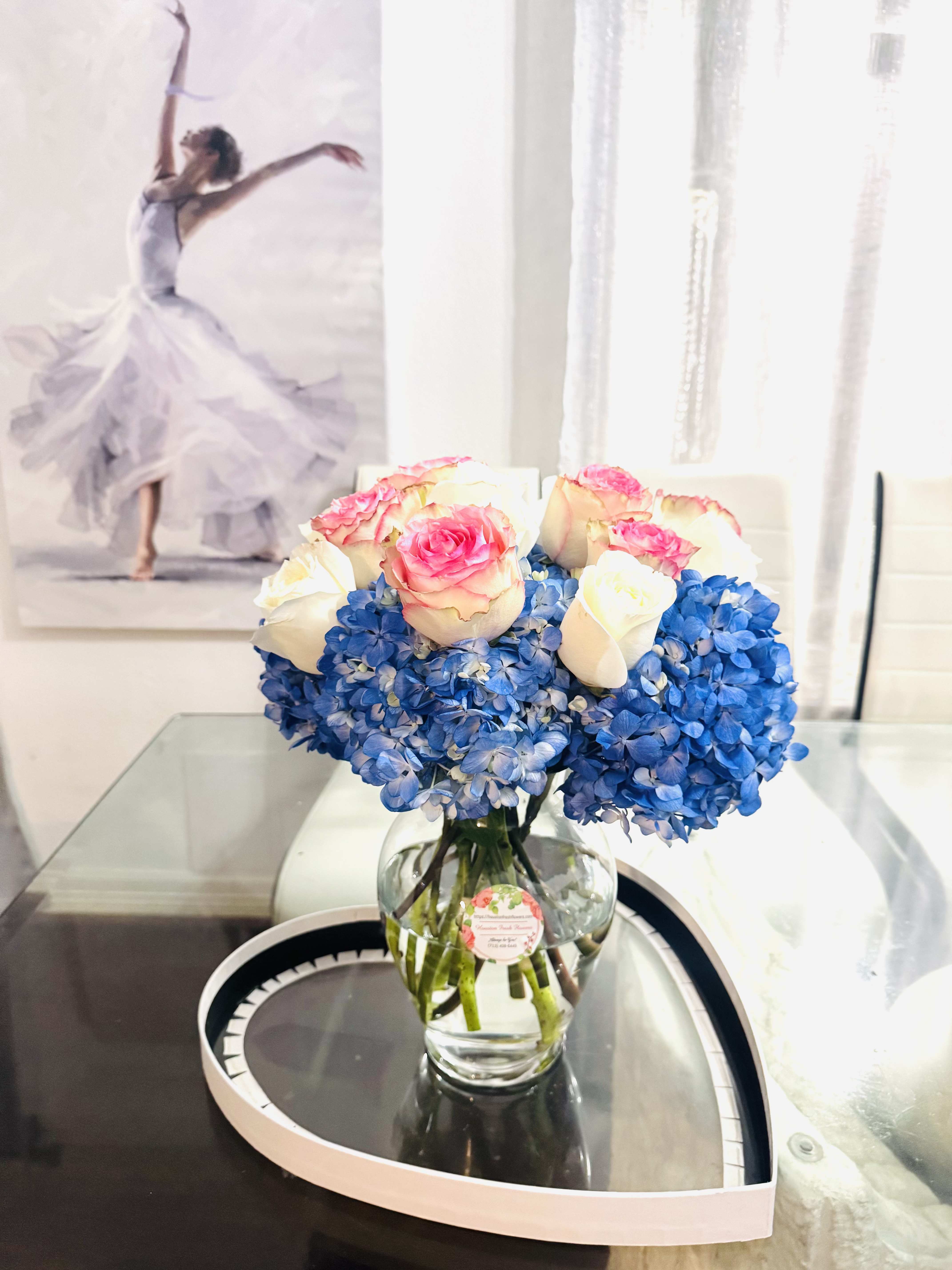 Ocean Kiss - This bouquet is refined and graceful, with a serene balance between freshness and romance. The base is formed by lush, deep blue hydrangeas, creating a soft, cloud-like foundation that feels both calming and elegant. Rising above are delicate roses in creamy white and blush-pink tones, with the pink edges glowing like watercolor brushstroke. The glass vase enhances the clean, contemporary look. The overall impression is that of romantic purity with a modern touch. 