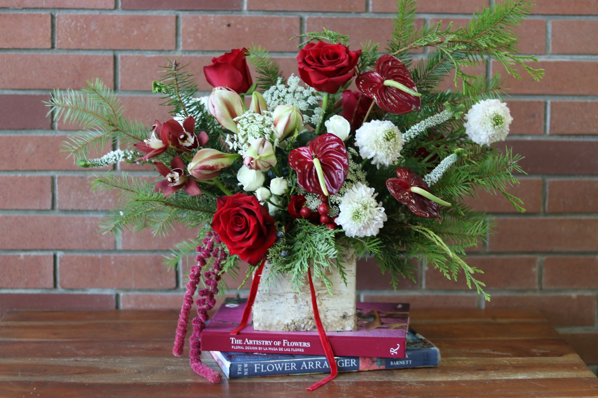 Crimson Winter Elegance  - **Name:** &quot;Crimson Winter Elegance&quot;  **Description:**   This stunning modern flower arrangement centers around a sleek birch square vase, exuding a natural yet contemporary charm. The bold red anthuriums and vibrant red roses create a passionate foundation, while delicate scabiosa add a touch of whimsy. The striking candy cane amaryllis bursts forth with its unique striped blooms, harmonizing beautifully with the intricate textures of Queen Anne’s lace.   Adding an exotic flair, the brilliant red cymbidium orchids lend an air of sophistication, contrasted by the soft white Veronica that provides a light, airy quality. To complete the arrangement, mixed evergreens weave through the design, bringing a refreshing and festive touch. &quot;Crimson Winter Elegance&quot; is perfect for those seeking to infuse their space with warmth and modern beauty, making it an ideal centerpiece for any occasion.