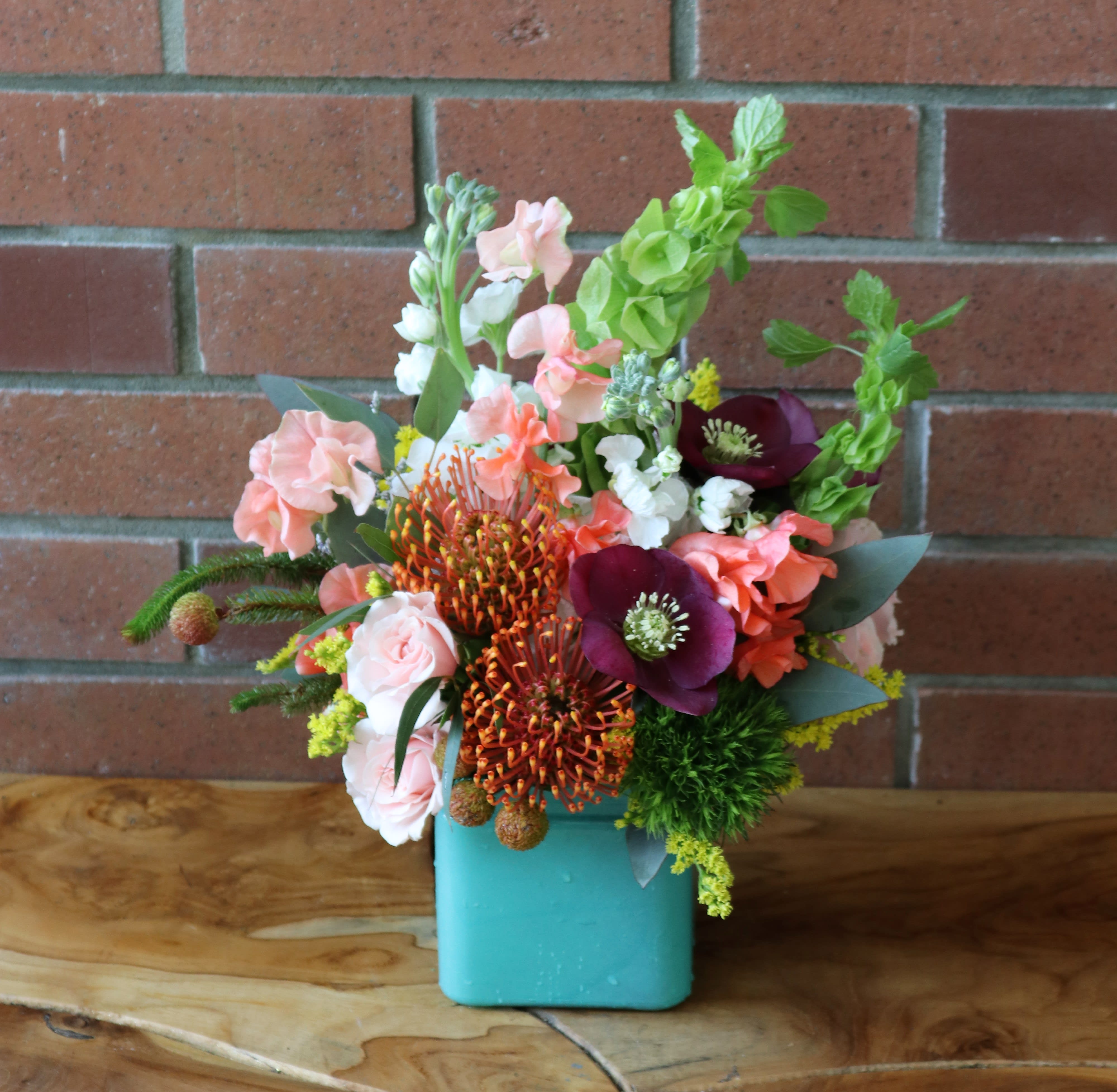 Charming - Coral Sweet Peas, beautiful bells of Ireland, tropical pin cushon proteas, helleborus, spray roses, trick and more in a blue cube vase. Perfect for any occasion!