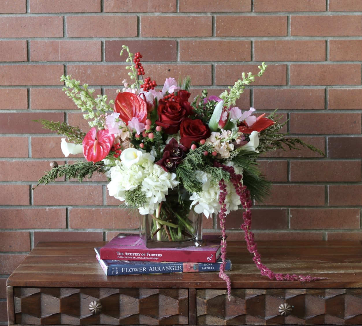 Christmas Dreamscape  - The &quot;Christmas Dreamscape&quot; arrangement is a breathtaking fusion of luxury and seasonal beauty, artfully presented in a modern cube vase. This exquisite design features lush hydrangeas and rich evergreens, creating a stunning foundation that embodies the essence of winter.   Bold anthurium adds a striking focal point, while trailing red amaranthus introduces a playful, cascading element. The arrangement is further enhanced by vibrant red roses, elegant white larkspur, and delicate pink sweet peas, all contributing to a harmonious blend of colors and textures.   White calla lilies and tulips add a touch of sophistication, elevating the overall elegance of the bouquet. The combination of these elements creates a captivating visual that evokes a sense of wonder and enchantment, making &quot;Christmas Dreamscape&quot; a perfect centerpiece for festive gatherings or a cherished gift that embodies the magic of the holiday season.