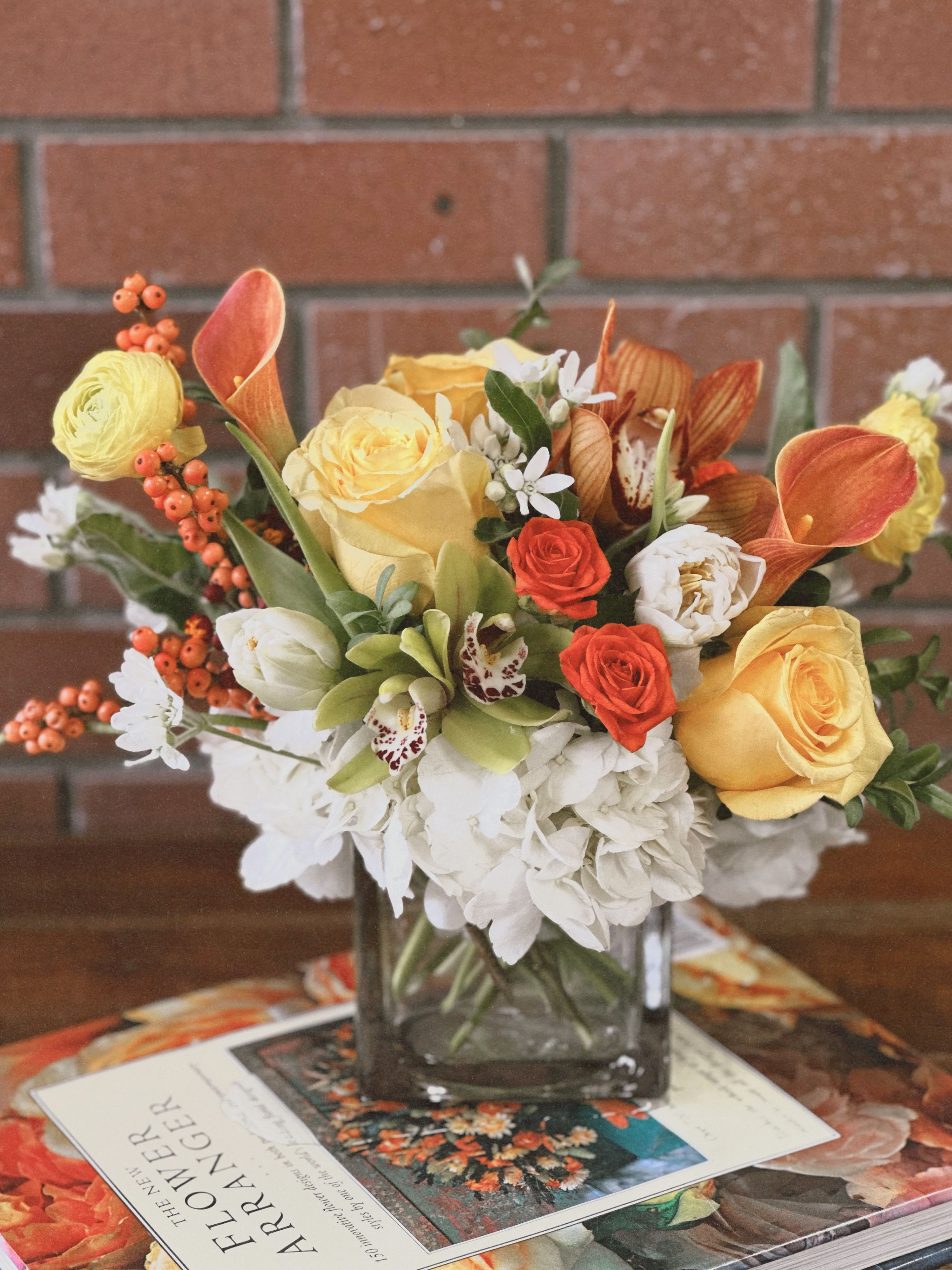 Harvest Harmony  - Envision a modern fall arrangement elegantly displayed in a sleek square cube vase. The design features lush hydrangeas in soft, muted hues, creating a plush foundation for the vibrant pops of color. Mini calla lilies, with their graceful curves, add an artistic touch, their creamy tones contrasting beautifully with the surrounding flowers.  Bright yellow roses bring a sense of cheerfulness, infusing the arrangement with warmth and brightness. The unique green cymbidium orchids introduce a fresh, contemporary element, their rich color providing a striking contrast to the oranges and yellows.  Scattered throughout, clusters of orange ilex berries add a playful, textural accent, while the cheerful yellow ranunculus introduce a sense of whimsy and fullness. Premium filler flowers enhance the composition, tying the colors together harmoniously. The overall palette of orange and yellow, punctuated by vibrant greens, creates a stunning focal point that embodies the essence of modern fall elegance.