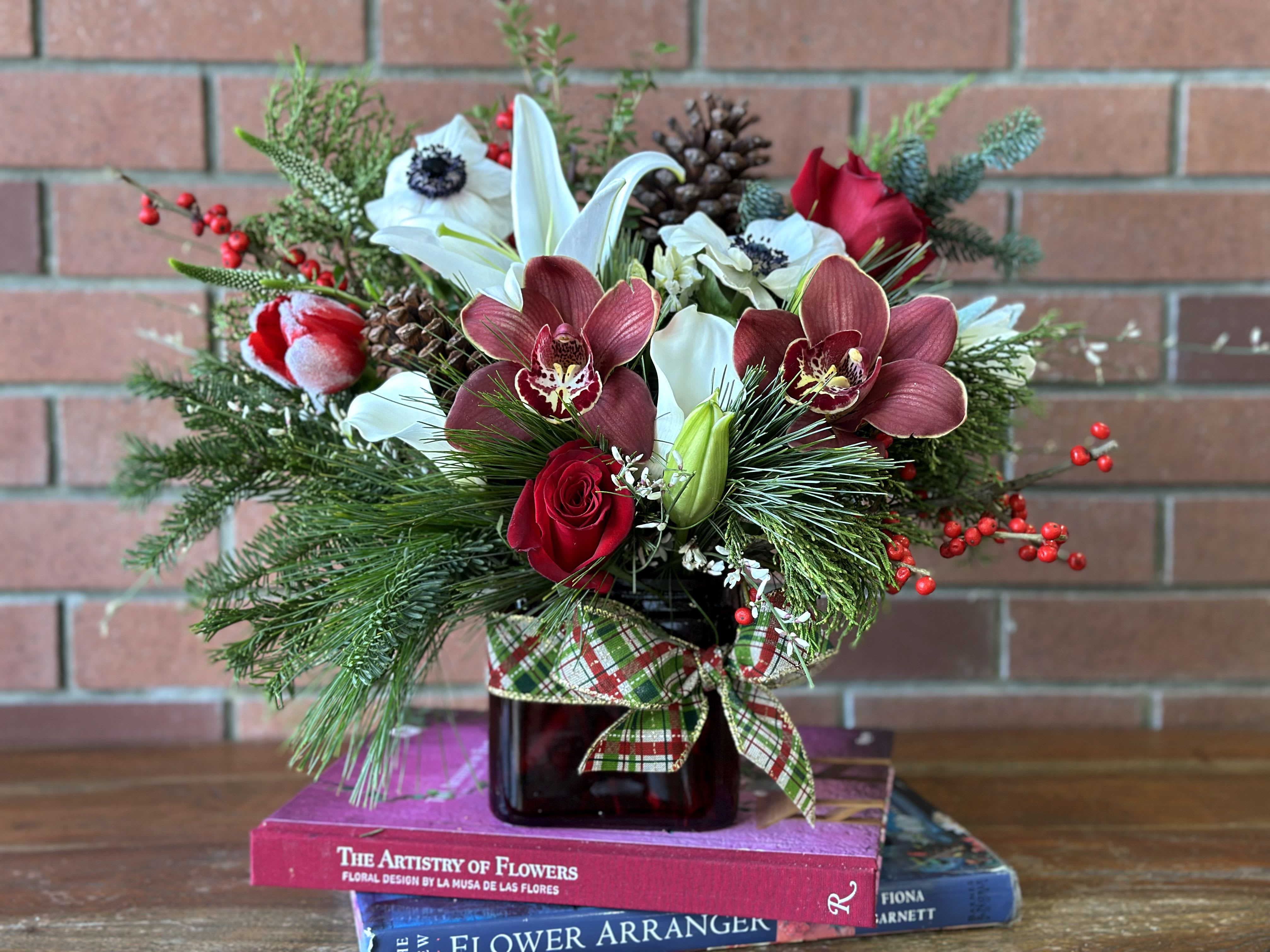 Winter Berry Bliss Bouquet  - Embrace the magic of the season with our &quot;Winter Berry Bliss&quot; arrangement. This stunning centerpiece features an exquisite blend of chocolate-colored cymbidium orchids, vibrant red roses, and pristine white lilies, all artfully arranged in a modern square vase. Accented with lush holiday ribbons, delicate filler flowers, and festive berries, this arrangement brings a touch of elegance and warmth to any holiday setting. The rich tones and textures create a captivating visual experience, perfect for enhancing your Christmas festivities or gifting to loved ones. Celebrate the beauty of winter with this sophisticated arrangement that embodies the spirit of the season!