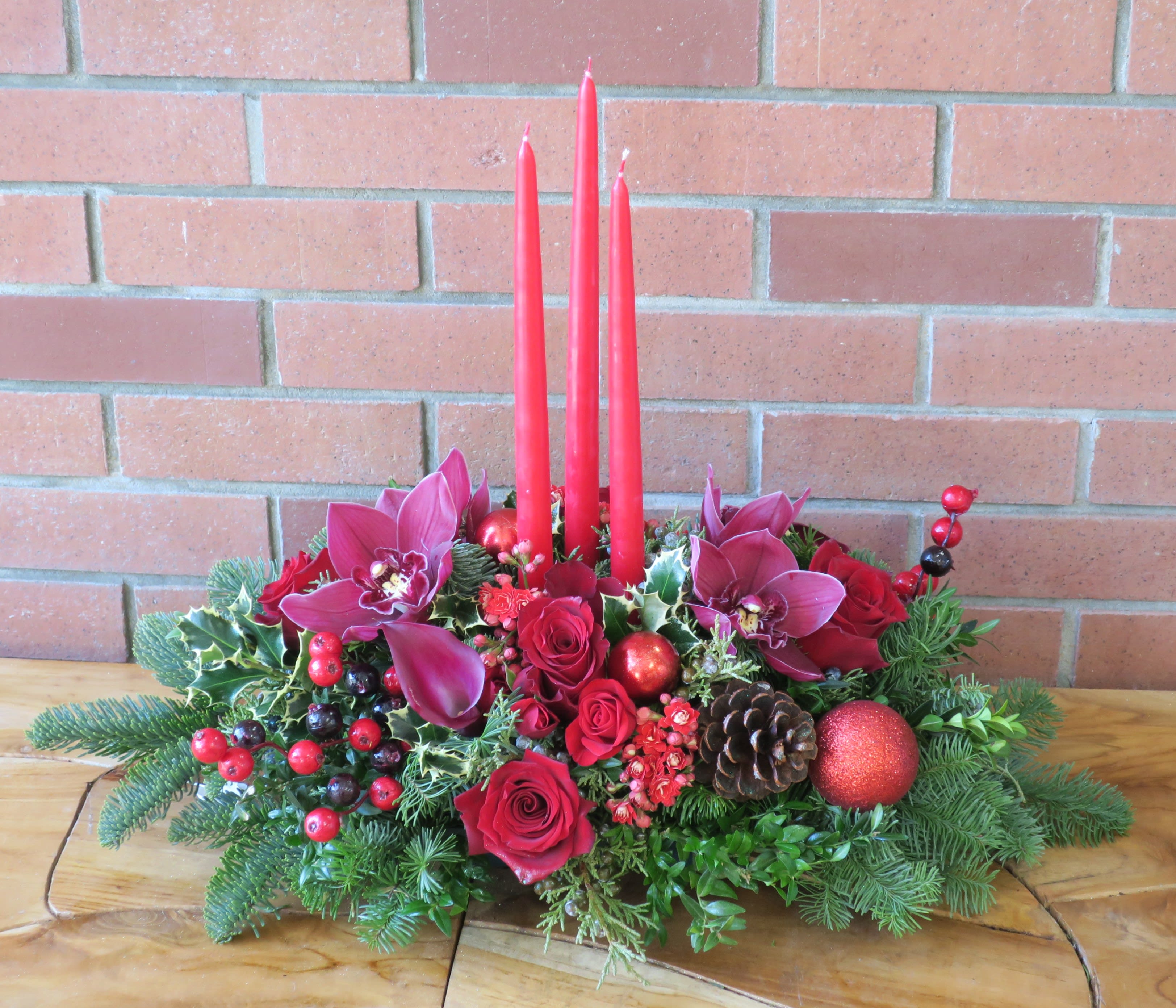 Tropical Traditions Centerpiece - Beautiful burgundy orchids, red roses, pine cones, evergreens and lush fillers with 3 red candles and berries. 