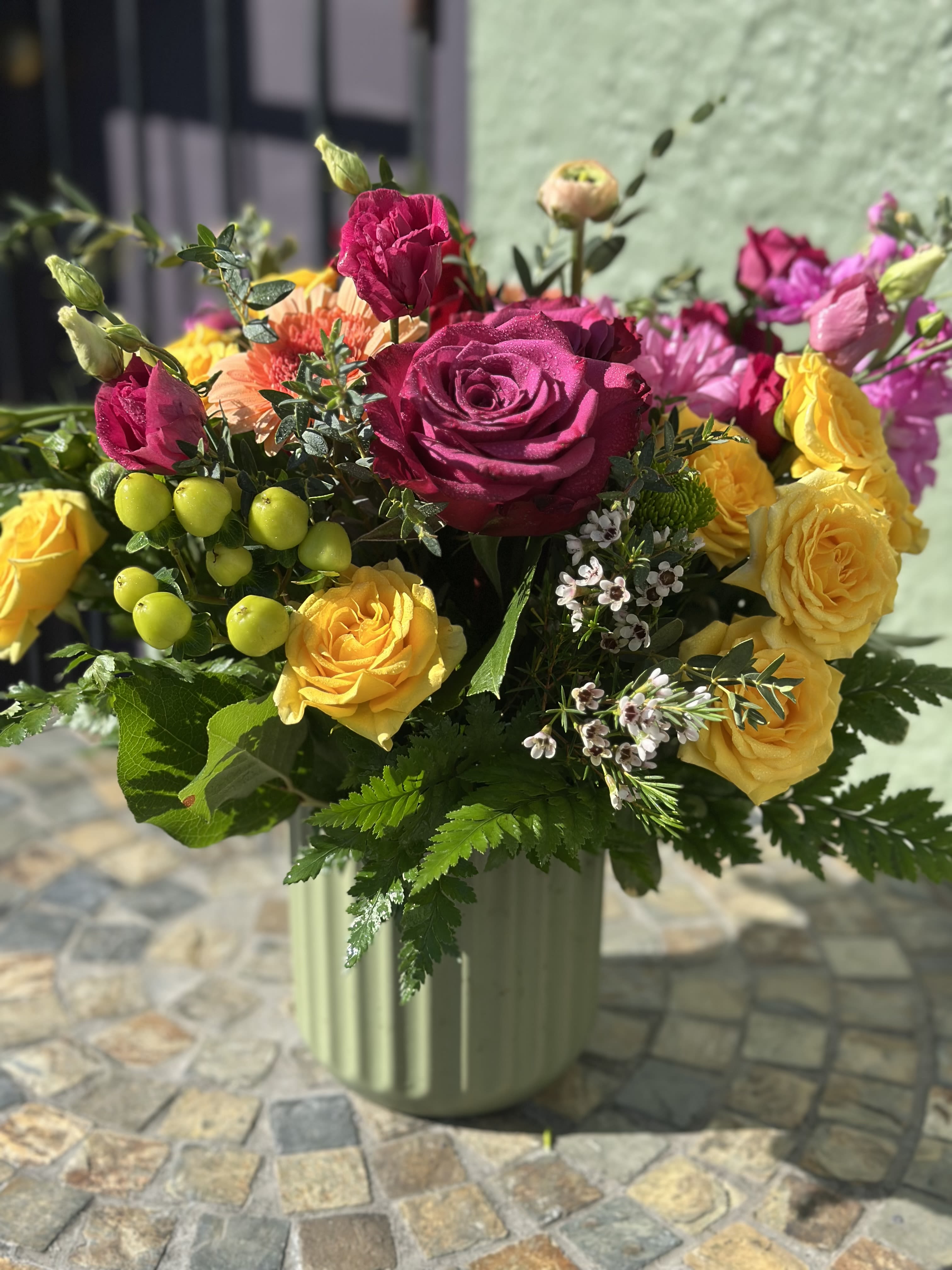 Sundara - Rich magenta roses mingle with golden yellow blooms, bursts of coral and soft pinks, and playful green accents — all gathered in a soft sage vessel that grounds the vibrant palette. It’s a celebration of color and light, perfect for brightening any space or mood.