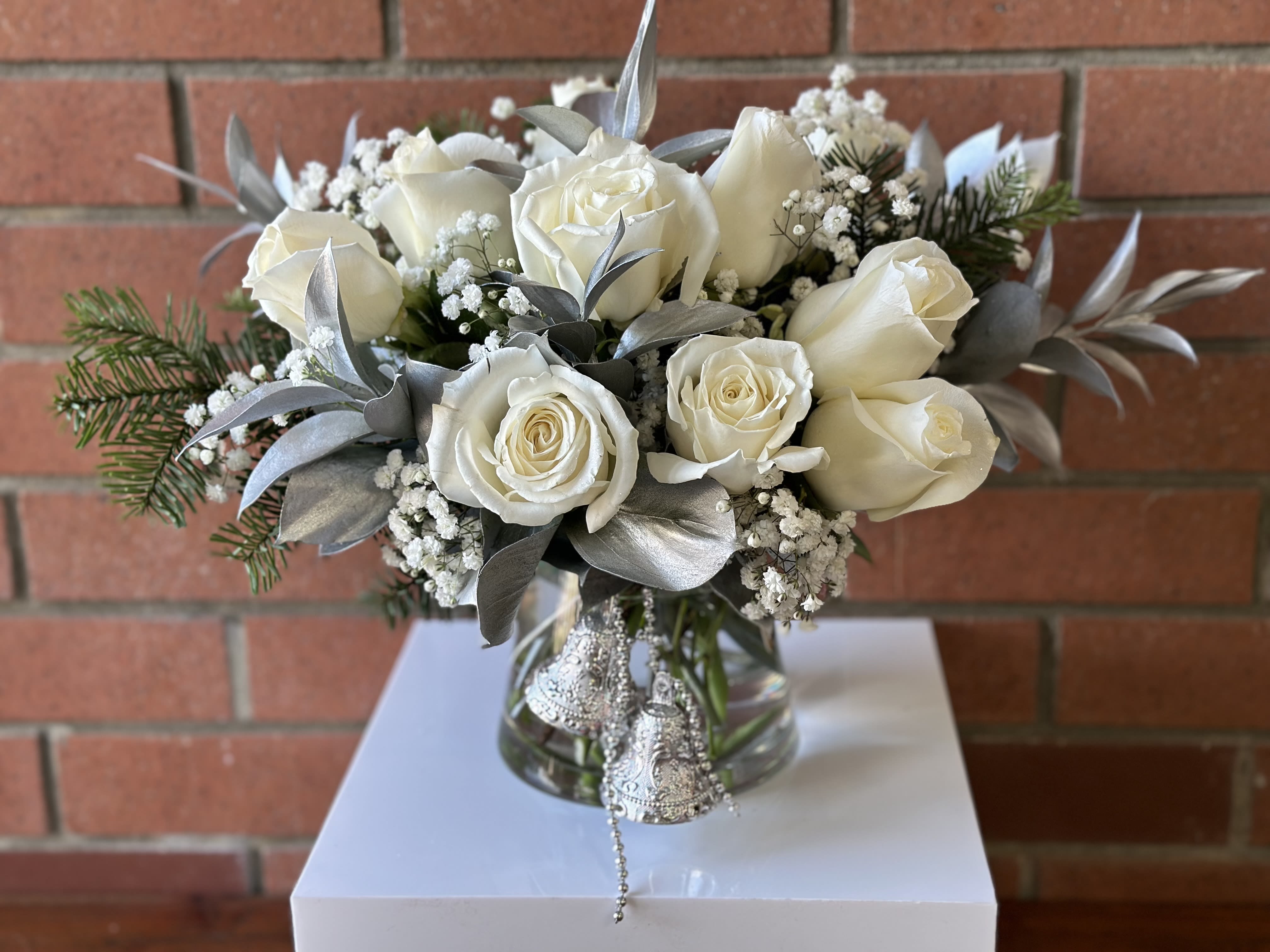 Silver Bells Roses  - White roses silver ruscus touches of fresh evergreens spruce/fir  baby’s breath in a modern clear glass vase.  
