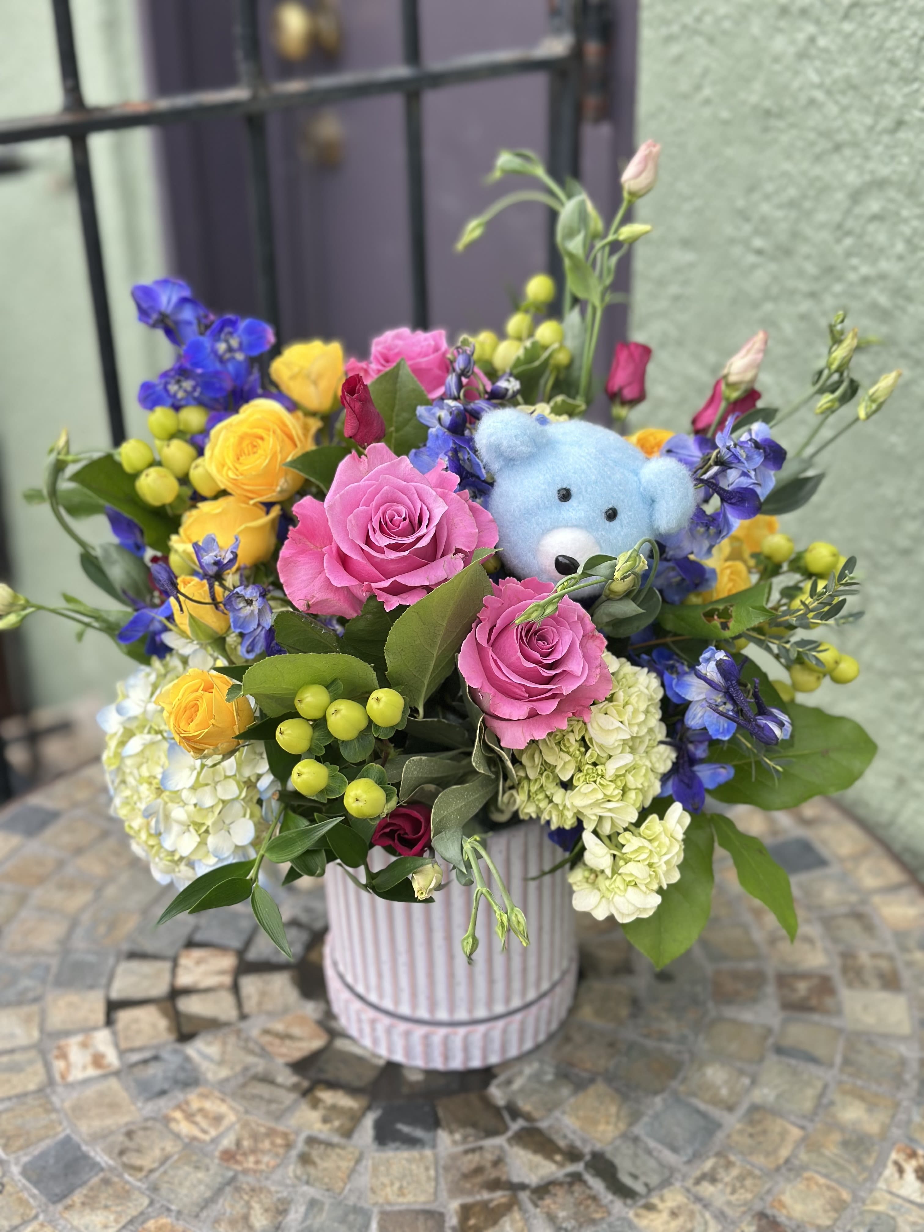 Oliver - Welcome the precious new arrival, &quot;Oliver&quot;, with this vibrant and playful arrangement. This design bursts with cheerful yellow roses, soft pink roses, and elegant white and blue hydrangeas and delphiniums. A sweet blue plush teddy bear pick (only head)is tucked among the blooms, adding cuteness. The bright colors are a lovely and cheerful alternative to traditional blue blooms, spreading joy and warmth to celebrate this special milestone.