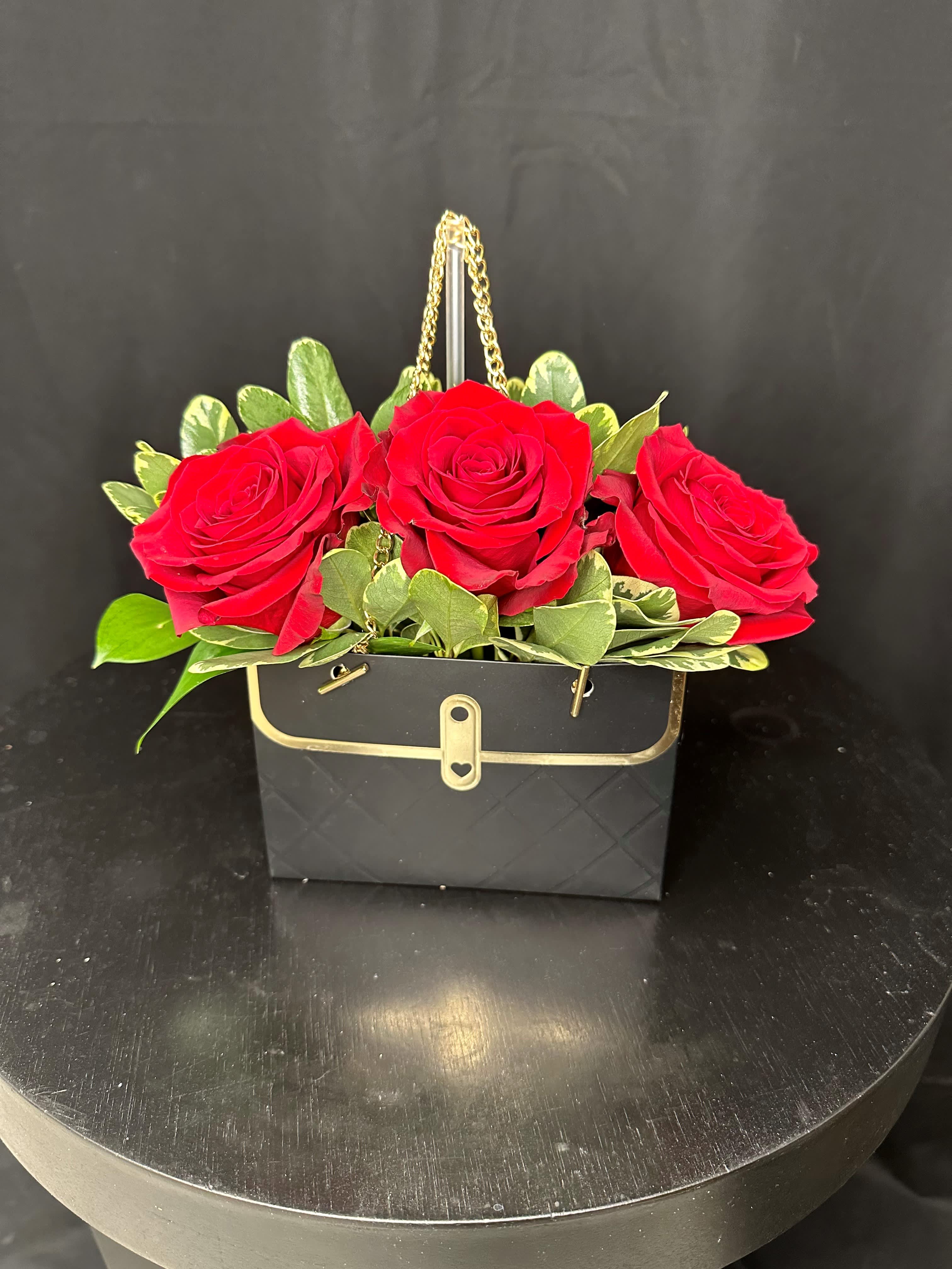 Eva -  Make her day with a captivating surprise - an arrangement of six striking red roses presented in a stunning black purse-shaped container adorned with luxurious gold accents. This elegant ensemble is sure to leave her speechless and feeling cherished. Let the beauty of these roses convey your love and appreciation in a memorable and stylish way.