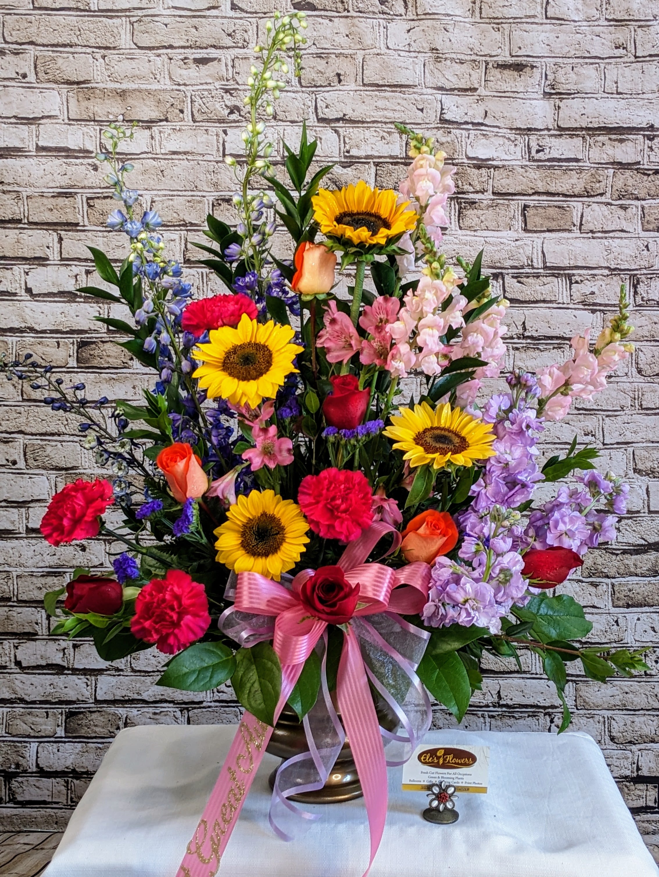 Garden of Memories - Bright and heartfelt, this tall sympathy design brings together a vibrant mix of sunflowers, roses, stock, snapdragons, carnations, larkspur and delphinium in a lush, garden-style tribute. Bold pops of yellow, red, coral, lavender, and blue are arranged in a high, sweeping silhouette—symbolizing hope, love, and cherished memories that continue to bloom. Finished with a soft pink bow and sympathy ribbon, this arrangement offers comfort through color and the quiet beauty of nature’s embrace.  Blooms may vary from photo.  Approximate Dimensions: 37"H x 30"W