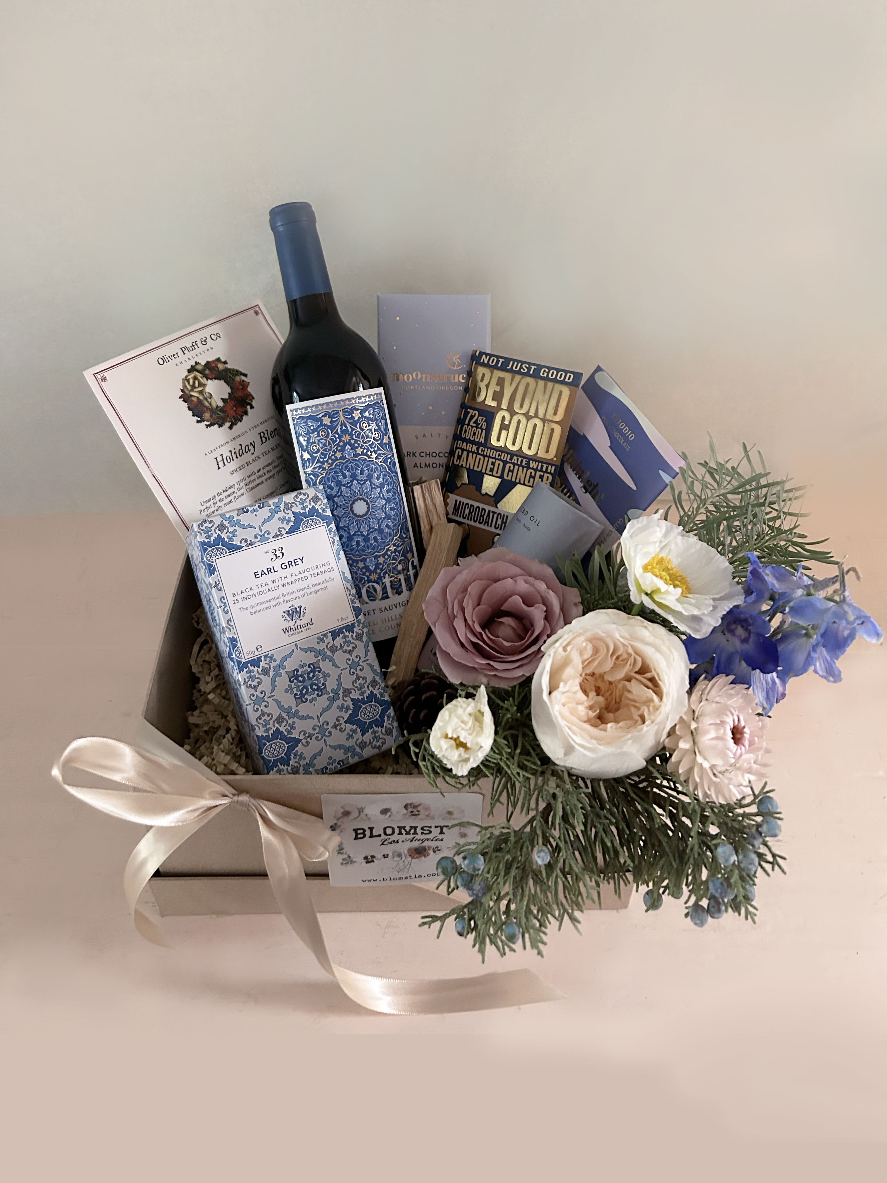 Happy Holidays Box - Everything needed for the cozy season in blue green and white colors. The box contains: Calming chamomile oil. Organic bath + body oil formulated to soothe muscles, joints, and skin, in and out of the tub. Perfect for those with sensitive skin with just a hint of sweet chamomile extract. Harney and Son's &quot;Winter White Earl Grey Tea&quot;  Sauvignon Blanc red wine. Holiday Tea blend, 6 teabags. Unwrap the holiday spirit with an aromatic blend of citrus and cinnamon. Perfect for the season, this festive black tea blend is spicy and naturally sweet.  Moonstruck Dark Chocolate Bar with Sea Salt and Almonds, 68% Cacao. Handcrafted superior  Goodio Midnight Sun white chocolate White chocolate topped with Arctic cloudberry crumble. Nothing shines brighter than the midnight sun. Taste the feeling of endless light! The Finnish company Goodio produces small-batch, “bean-to-bar” vegan chocolate, using the finest cacao beans sourced worldwide. Beyond Good Candied Ginger dark chocolate bar. Bright, zesty ginger pieces bring out the fruit-forward notes in this single-origin heirloom chocolate. Mini vase with winter arrangement. Ready for hand delivery in LA. For similar baskets for shipping nationwide, visit: www.blomstlosangeles.com