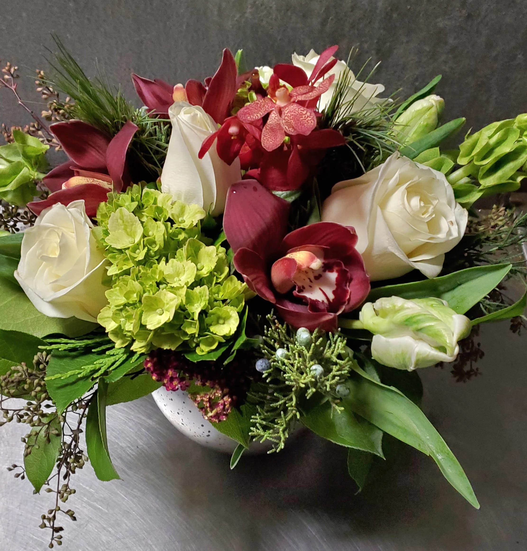 Burgundy Tidings   - Available Dec 9th on Burgundy and green are classic colors during this season, and they shine beautifully in this low  arrangement.  White roses are rimmed with mini green hydrangea, burgundy orchids and a variety of mixed greens that add the perfect touch of texture.