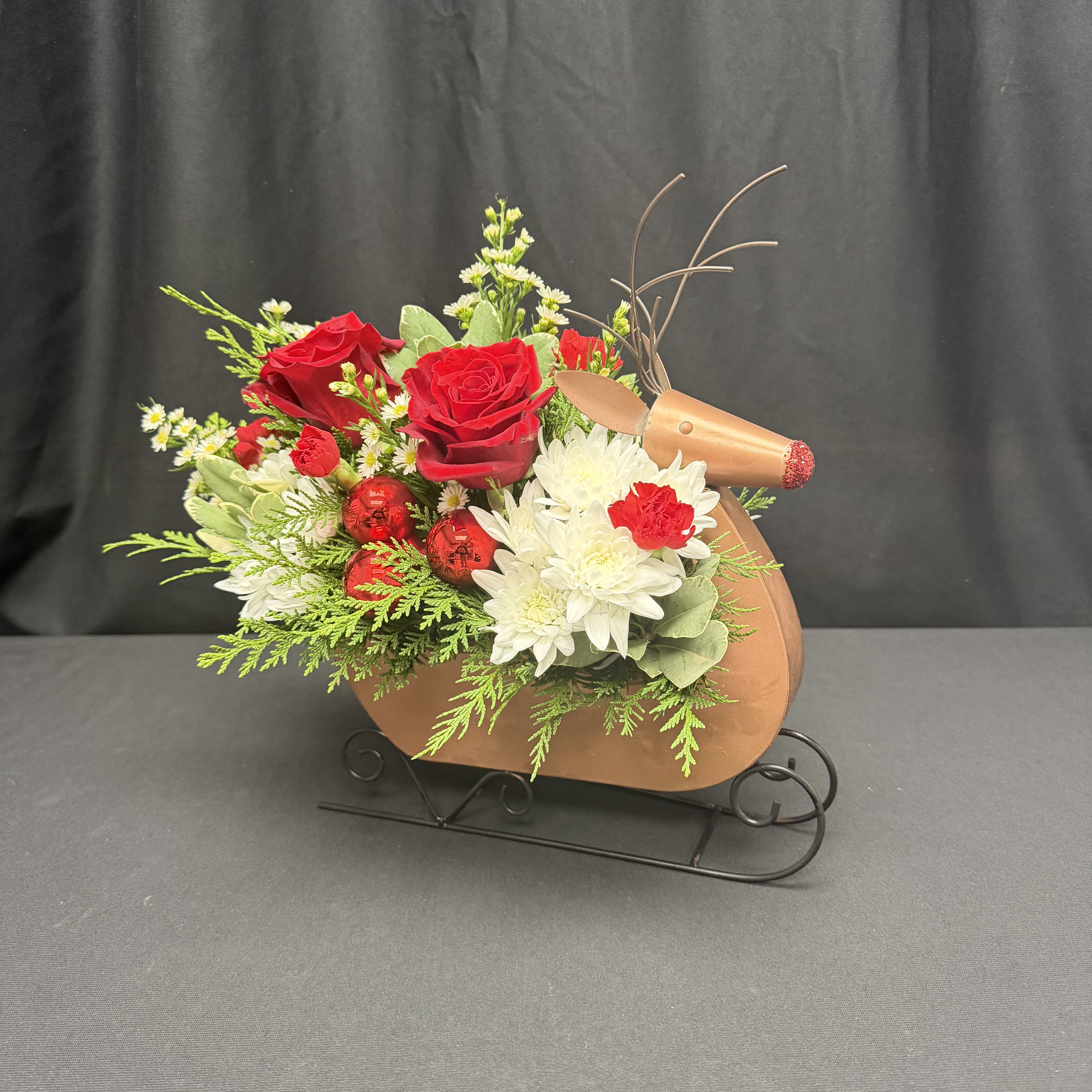Rudolph's Sleigh Ride - Bring a burst of Christmas cheer to your home with our charming North Pole Gnome arrangement. Nestled in an adorable holiday gnome container, this festive design features fluffy white hydrangea, vibrant red roses and carnations, and touches of winter greenery for the perfect pop of seasonal color. A tiny Santa hat adds a playful finishing touch, making this piece a joyful favorite for gifting or decorating.