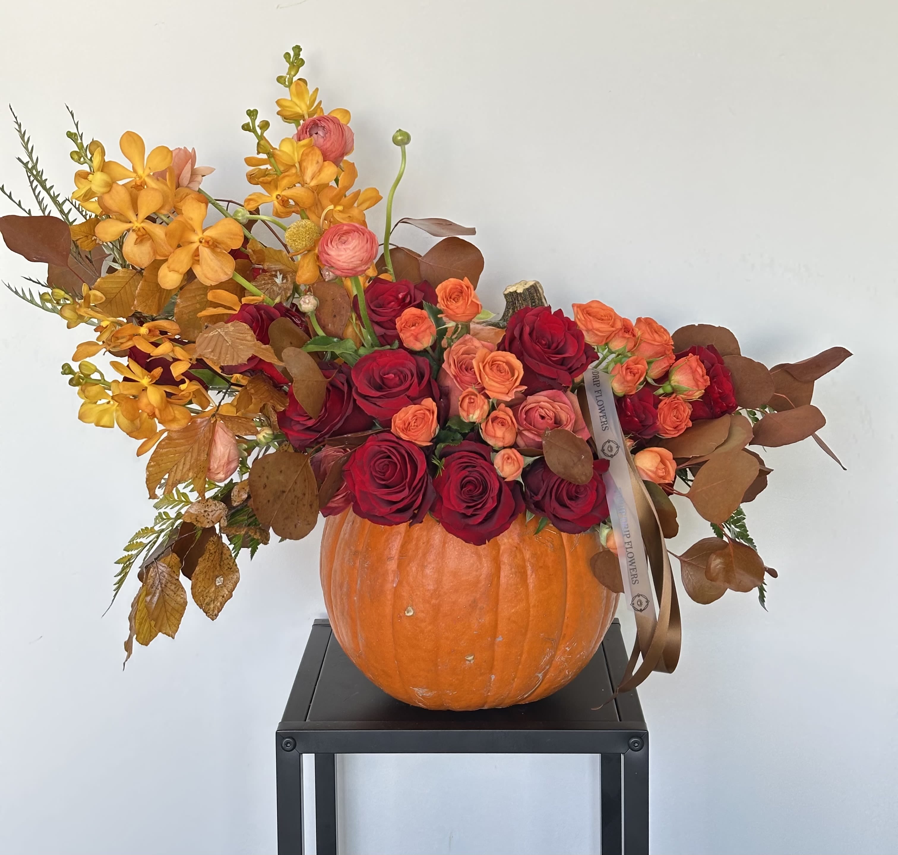 Autumn Isabella Pumpkin Arrangement - Celebrate the warmth of the season with our Autumn Isabella Pumpkin Arrangement, a stunning fall-inspired floral design crafted inside a real pumpkin. This luxurious piece features a rich blend of Roses, Spray Roses, Ranunculus, Orchids, and seasonal fall foliage, creating a vibrant mix of deep reds, oranges, and golden tones.  Perfect for Thanksgiving centerpieces, fall birthdays, holiday dinners, housewarmings, or seasonal home décor, this arrangement brings a cozy, elegant harvest feel to any space. Designed exclusively by TulipDripFlowers, it’s a show-stopping piece for anyone who loves autumn colors and unique floral art.