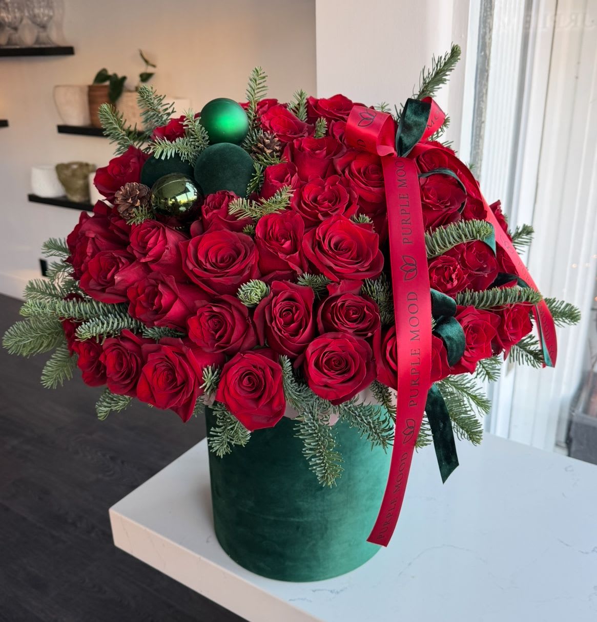 Holiday roses in Box - This is a luxurious arrangement of vibrant red roses, accented with fresh evergreens, pinecones, and festive ornaments. Designed in a plush green box and finished with elegant ribbons, this masterpiece captures the essence of Christmas joy and sophistication. Perfect for making a bold statement this holiday season, it’s an ideal gift or centerpiece for any festive gathering. 