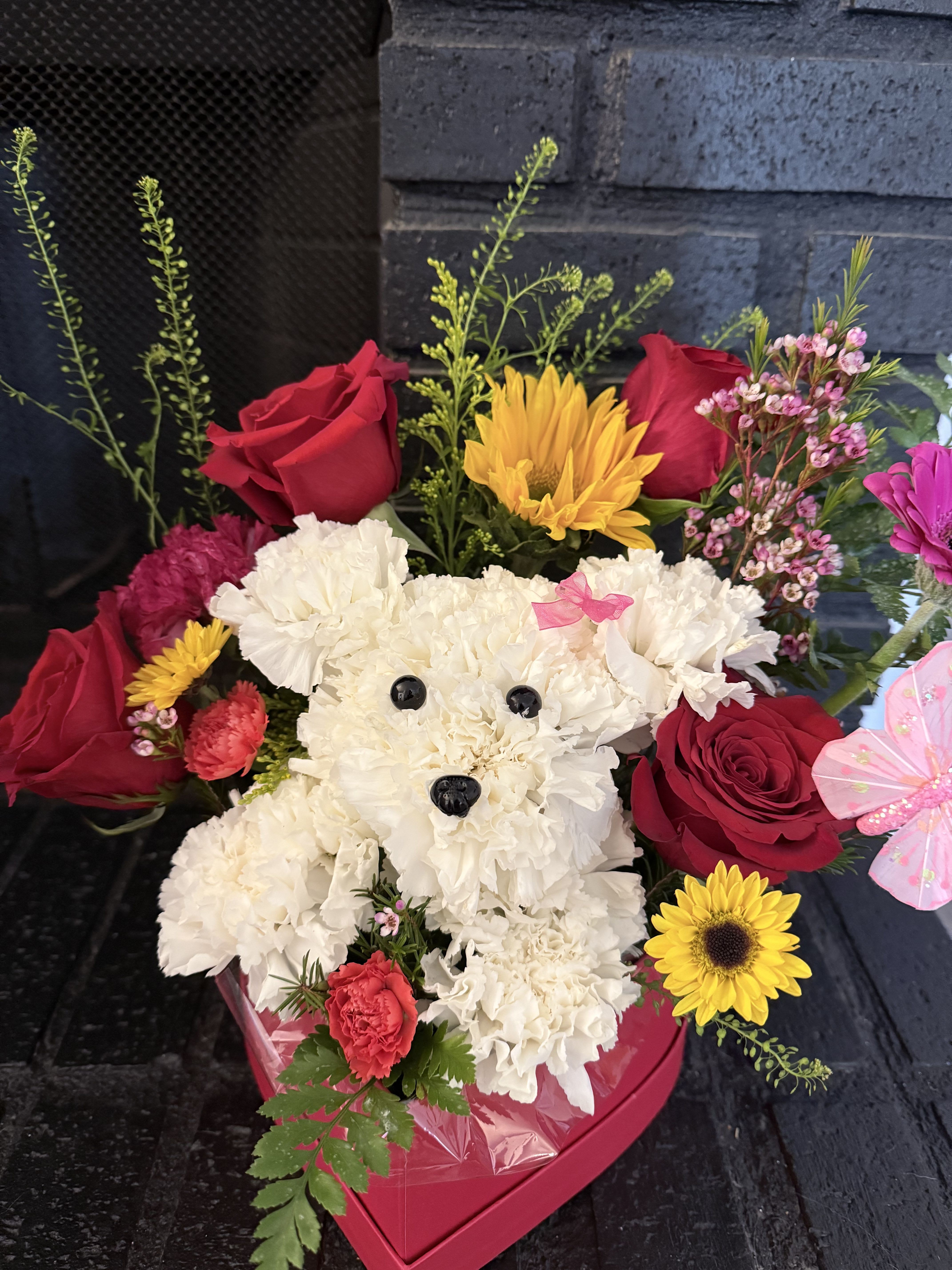 Floral Puppy Surprise - This charming arrangement features an adorable puppy handcrafted from fresh white carnations, surrounded by vibrant roses, cheerful sunflowers, mini carnations, and delicate seasonal blooms. Presented in a heart-shaped box, it’s the perfect gift to brighten someone’s day with sweetness, color, and love.