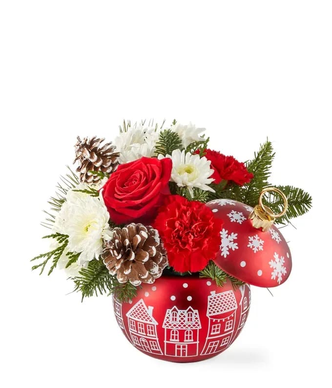 Winter Village Charm - Send a holiday gift that captures the warmth and charm of the season. This festive arrangement features lush red roses, bright red carnations, and soft white poms nestled in seasonal greenery, accented with rustic pinecones. Arranged in a red ornament-shaped container adorned with a white village scene and snowflake details on the lid, it brings cozy holiday magic to any space. Perfect as a cheerful centerpiece or thoughtful gift, this arrangement is a delightful way to spread joy and celebrate the beauty of the season.
