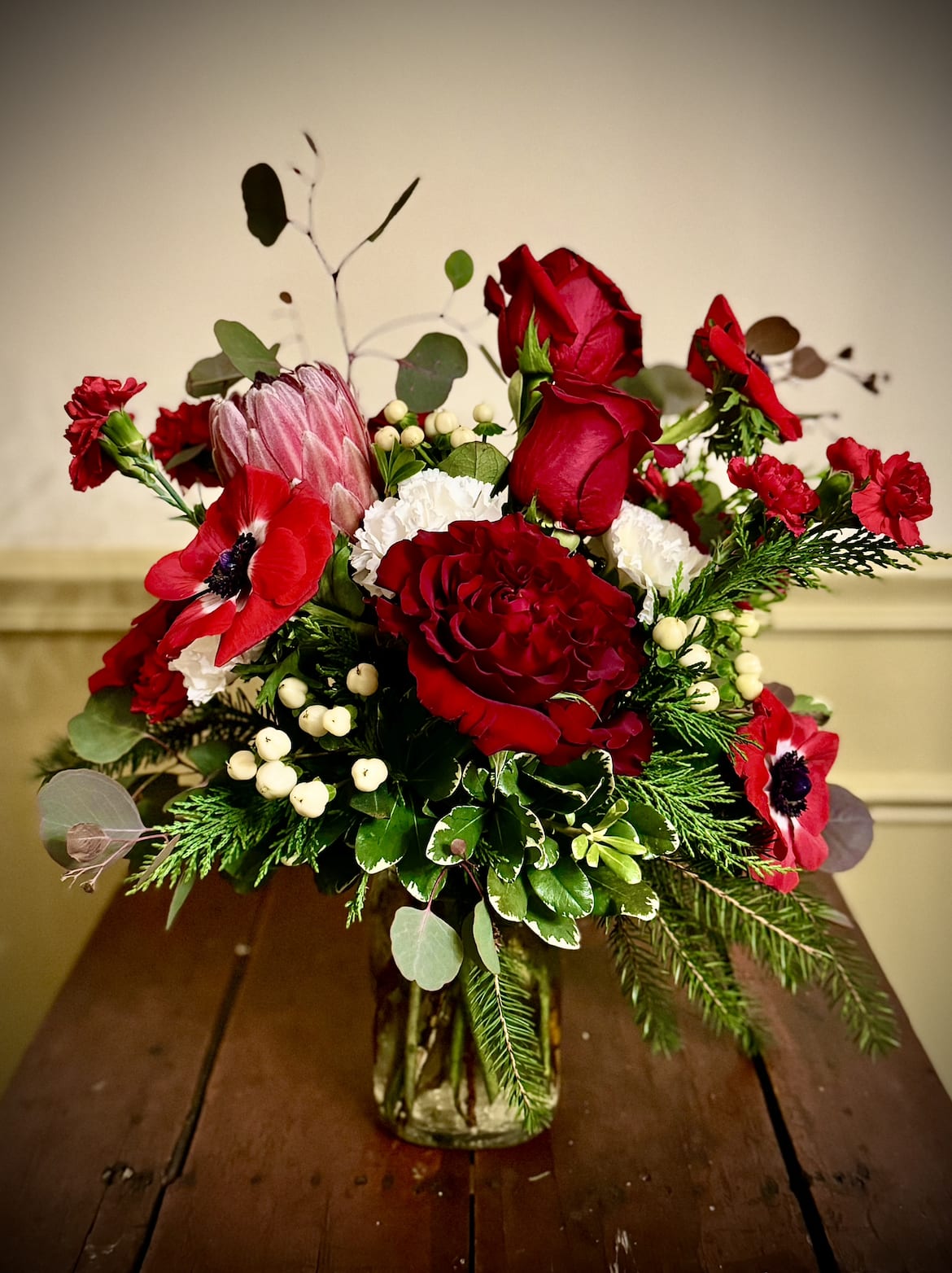 Christmas Magic - Embrace the magic of the season with this stunning arrangement of dark red roses, vibrant red anemones, exotic protea (with premium design), and lush seasonal greens. Designed in a classic glass jar.  Perfect for holiday elegance.  Perfect as a holiday hostess gift.