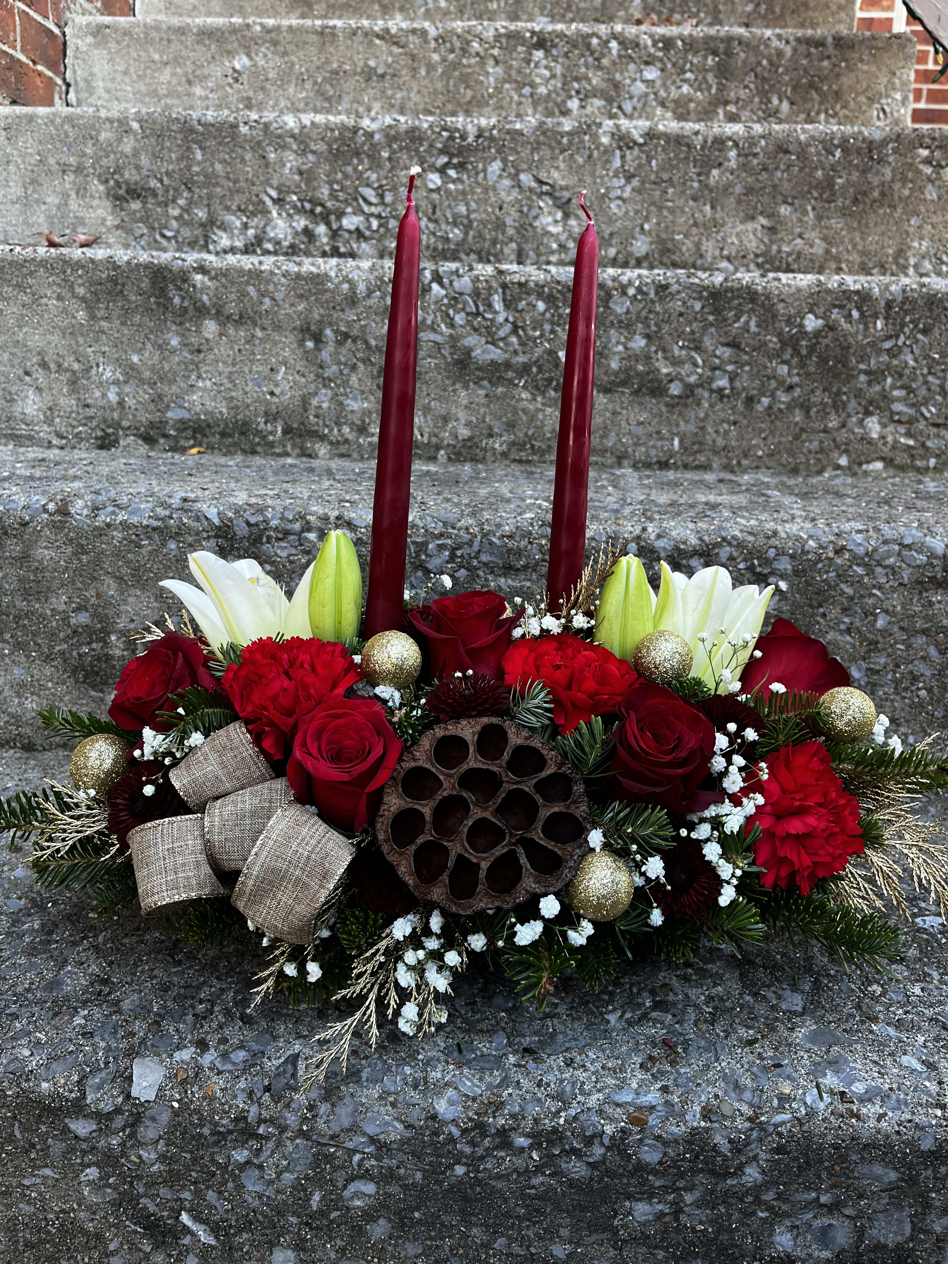 Santa’s Centerpiece  - This centerpiece has roses, lilies, pinecones, carnations, babies, breath, ribbon tuffs, Christmas ornaments, Christmas greens, Christmas greens, colored gold, Pod and two pomegranate colored candles.