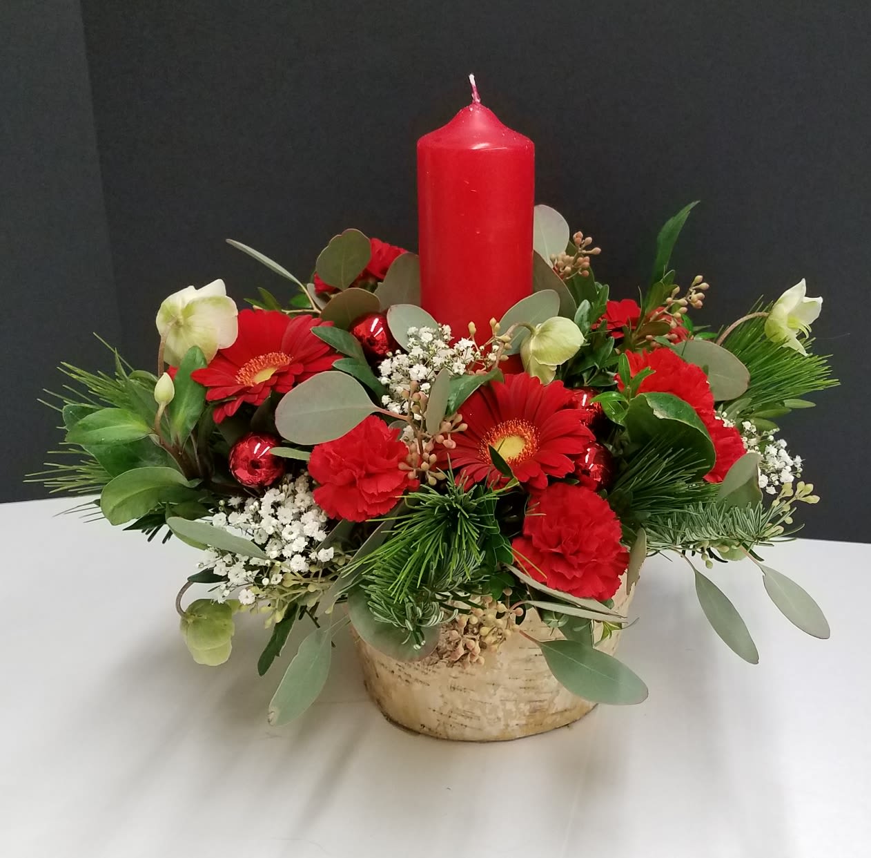 Christmas Glow Centerpiece  - A birch container with a red candle nestled in an assortment of winter greens, seeded eucalyptus, helleborus, baby's breath, red mini gerbera daisies, red mini carnations and finished with red ornament balls.