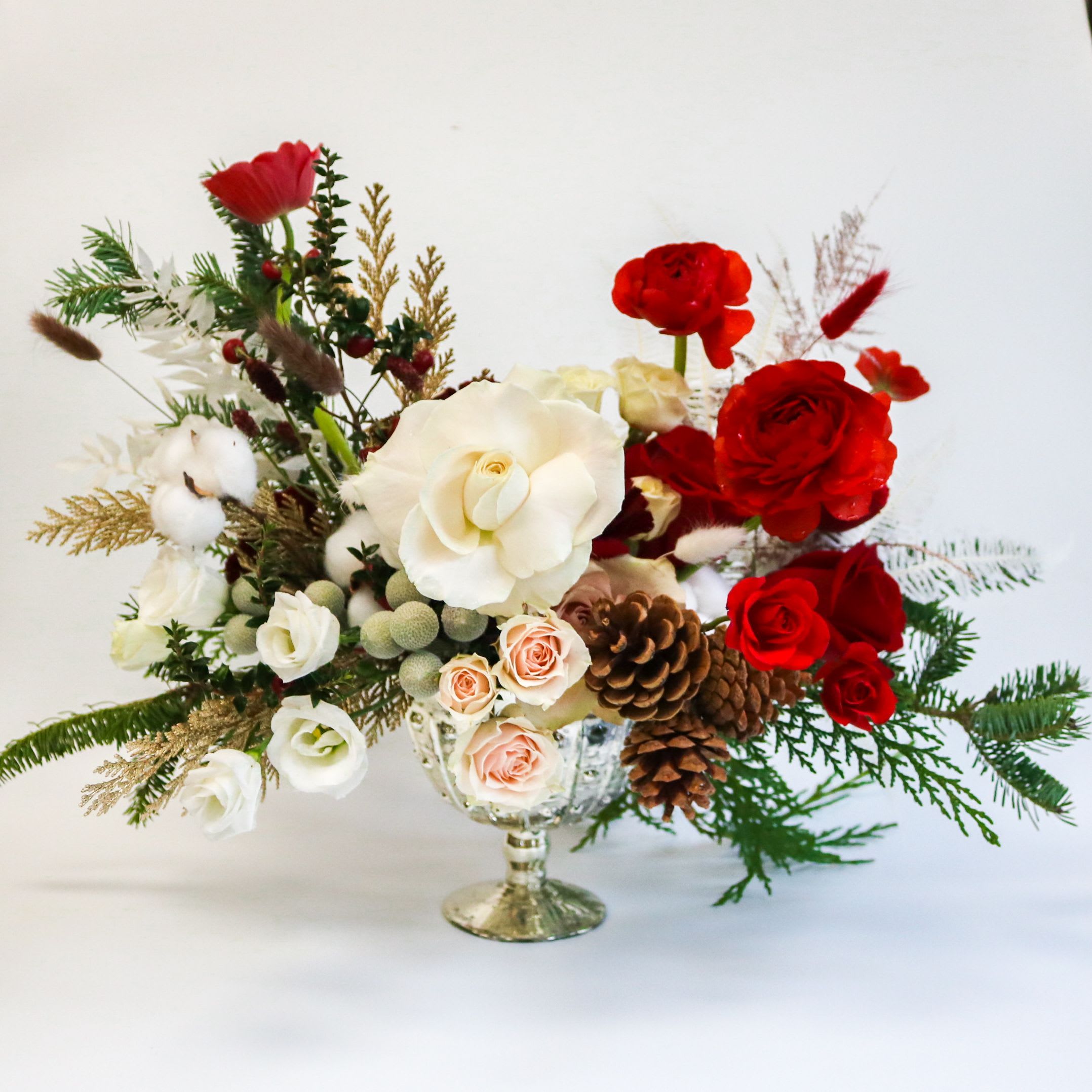 December Glow - A luxurious holiday centerpiece designed to bring warmth, elegance, and festive charm to your holiday table. Styled in a silver footed compote, this arrangement features a lush blend of premium blooms: velvety red roses, soft ivory garden roses, blush spray roses, and airy lisianthus.  Textural accents — cotton stems, pinecones, winter greenery, and golden foliage — add depth and seasonal magic, while delicate red touches provide the perfect pop of holiday color.  