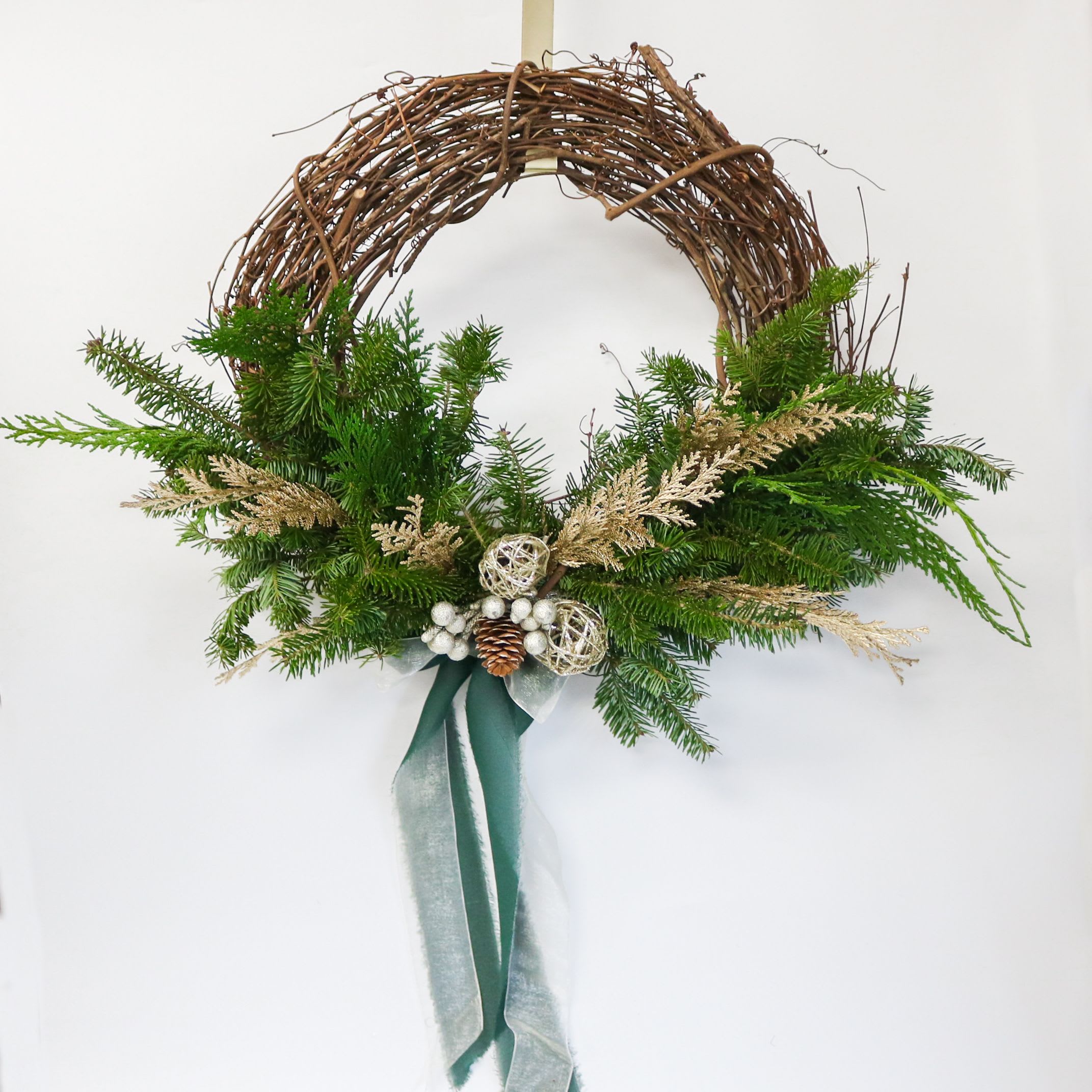 Noel Wreath 14in  - A modern, airy twist on the classic Christmas wreath — designed on an open grapevine base for a light, organic feel. The Evergreen Noel Wreath features a graceful sweep of fresh winter greens, mixed with soft golden foliage, frosted pinecones, and delicate ornament accents for just the right touch of holiday sparkle.  Hand-tied velvet and sheer ribbons in deep green and winter white add movement and elegance, making this wreath a beautiful statement piece for any home.  Handcrafted in the Bloomie studio, each wreath is unique, textural, and thoughtfully designed to bring understated holiday charm to doors, mantels, and entryways.